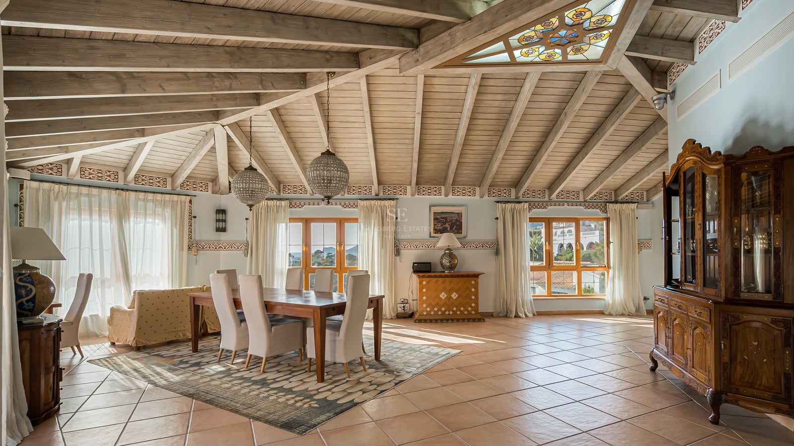 Salle à manger avec plafond voûté en bois, carreaux de terre cuite et grandes fenêtres lumineuses.