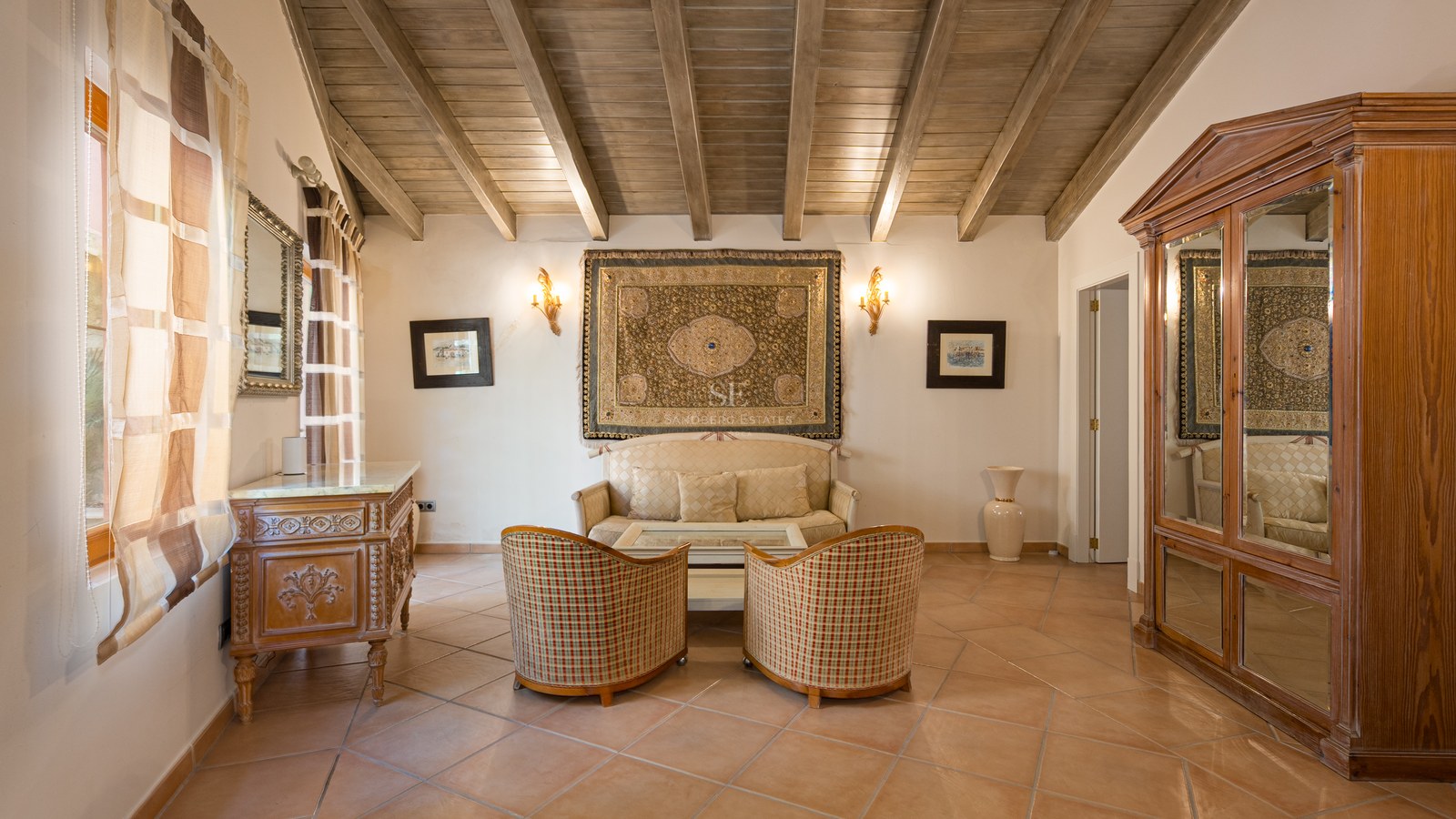 Espace de vie avec sol en terre cuite, plafond en poutres de bois, fauteuils en osier et tapisserie murale.