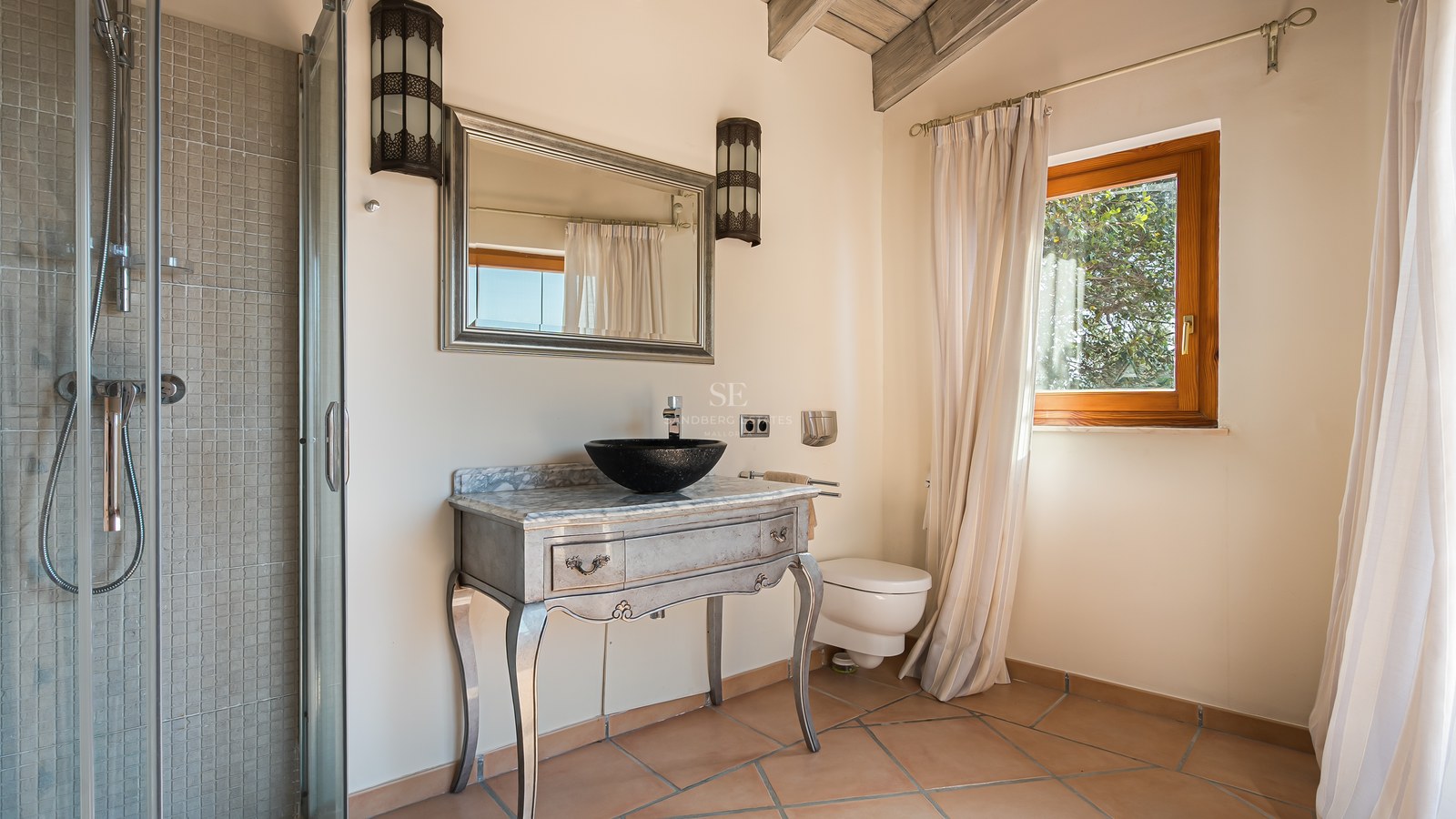 Salle de bains avec meuble argenté orné, vasque noire, sol en terre cuite et lumière naturelle.