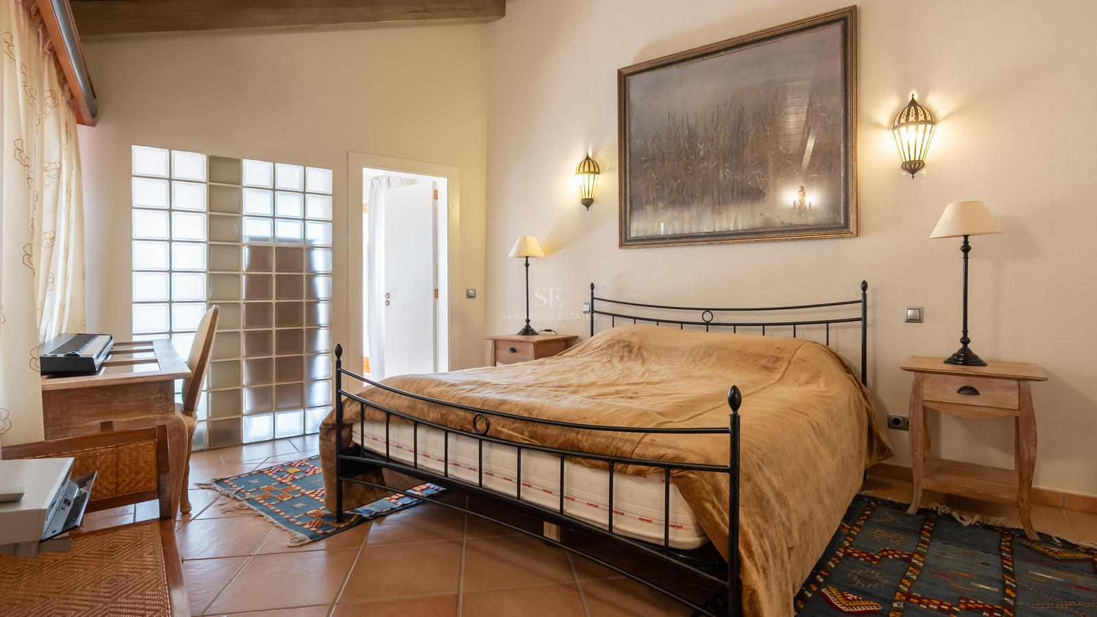 Chambre avec lit en fer forgé, sol en terre cuite et mur en briques de verre sous des poutres apparentes.