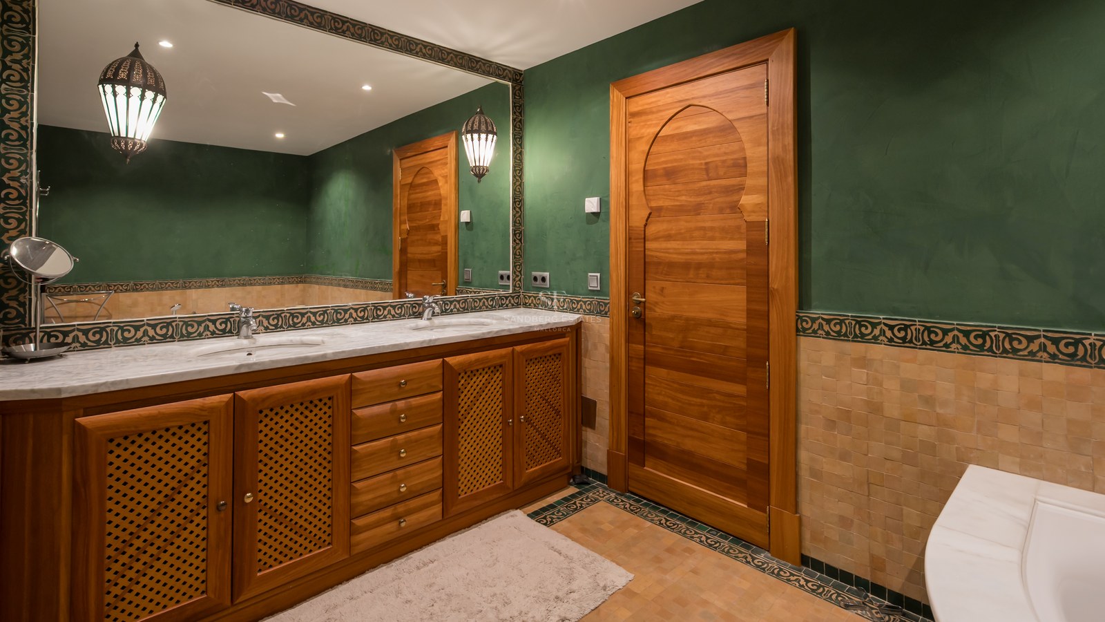 Salle de bain aux murs verts, meubles en bois sculpté, double vasque en marbre et grand miroir décoré.