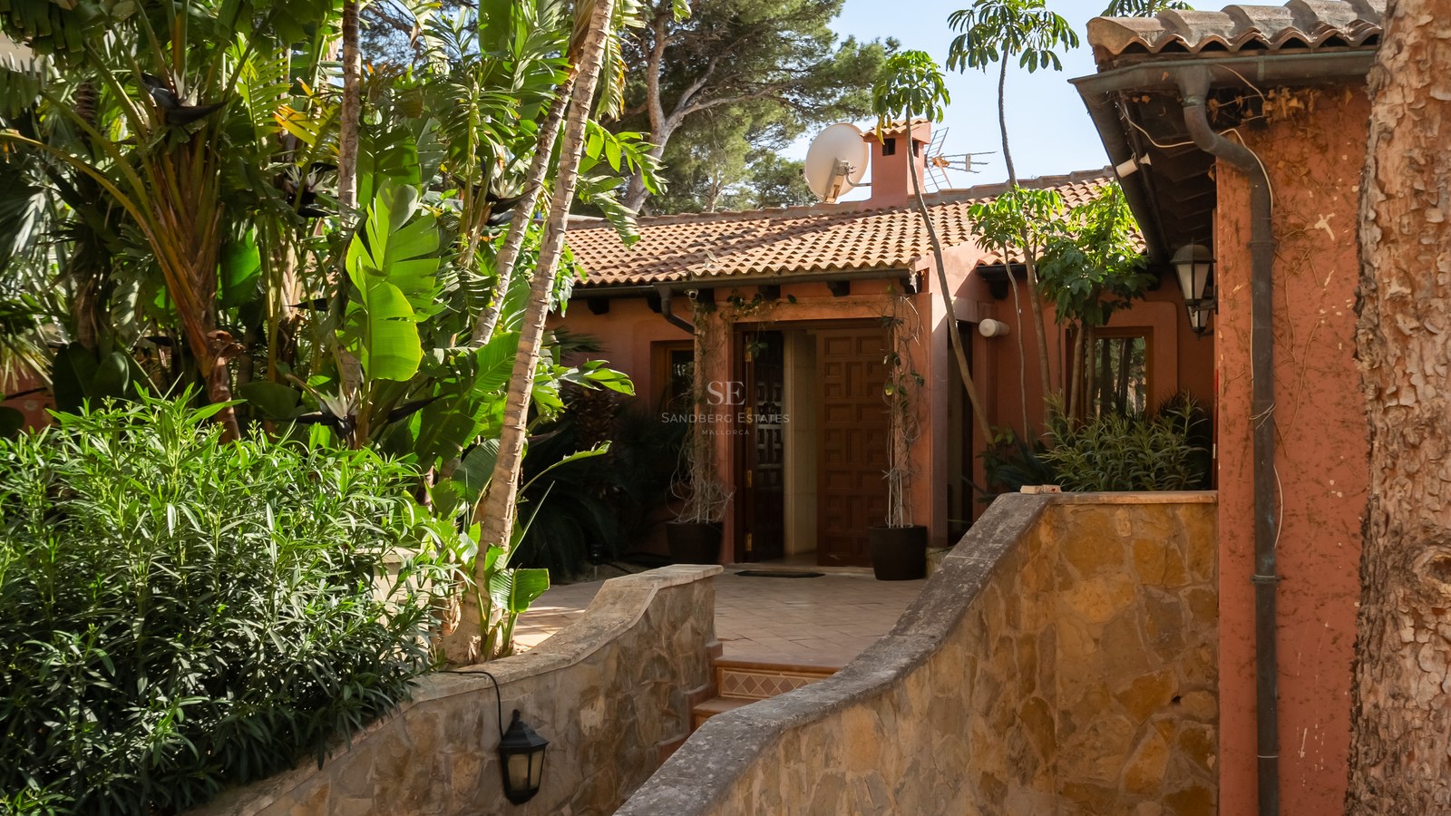 Entrée de villa aux tons chauds avec porte en bois, chemin en pierre et végétation tropicale sous un soleil radieux.