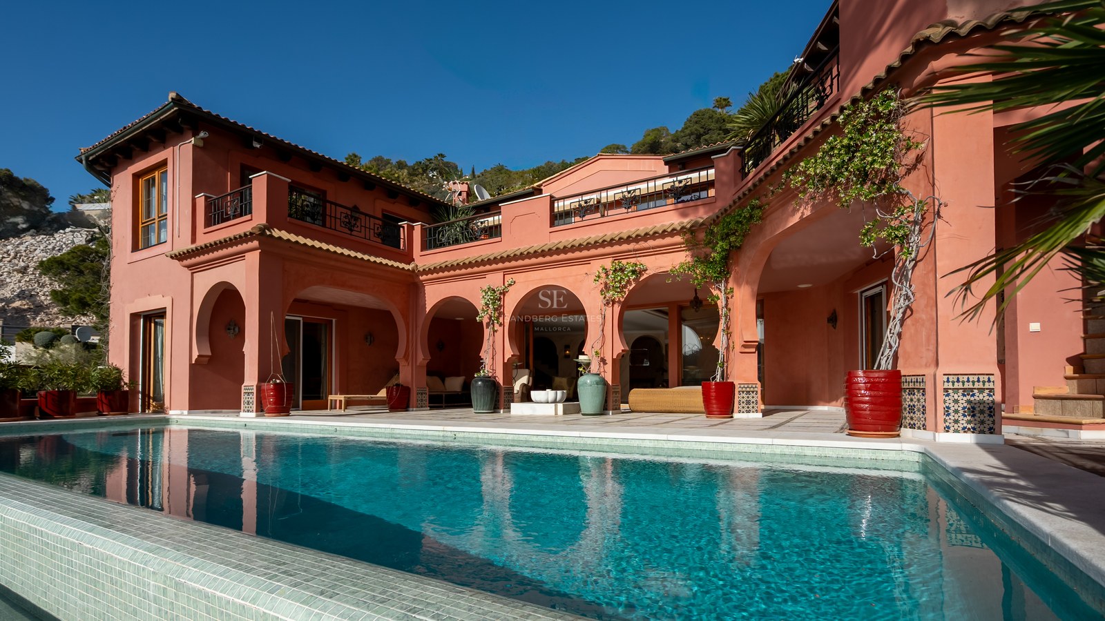 Grande piscine turquoise devant une villa terre cuite aux arches traditionnelles et terrasses ensoleillées.