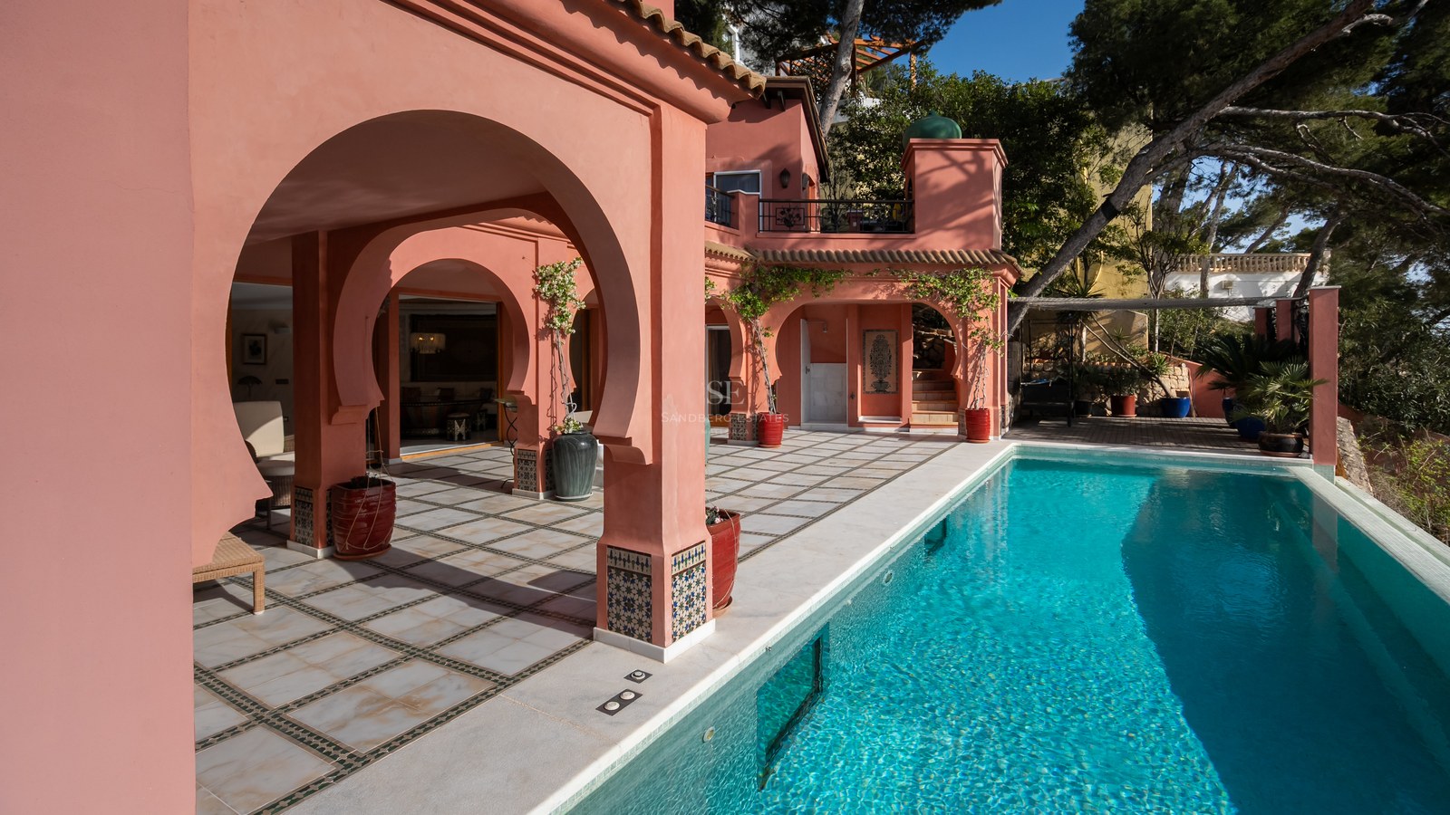 Piscine turquoise jouxtant une villa rose saumon avec arches mauresques et terrasse carrelée sous le soleil.