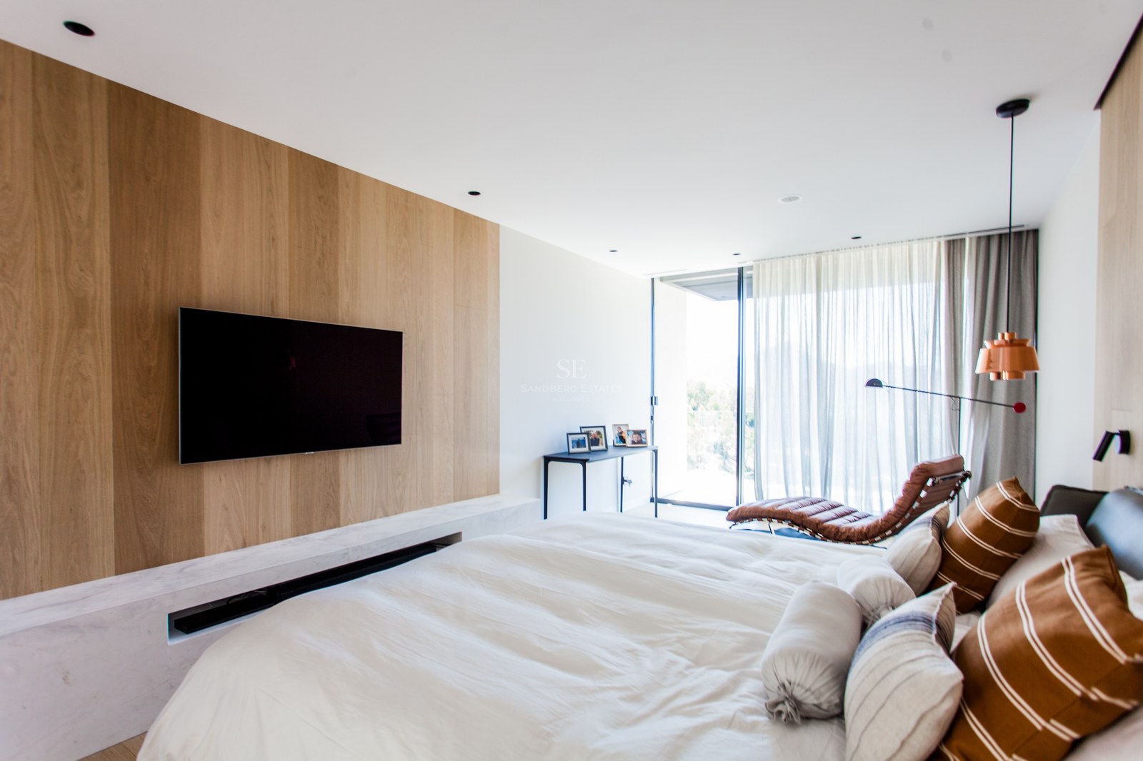 Modernes Schlafzimmer mit Eichenholz-TV-Wand, weißem Marmorsockel und Leder-Loungesessel vor Terrassentür.