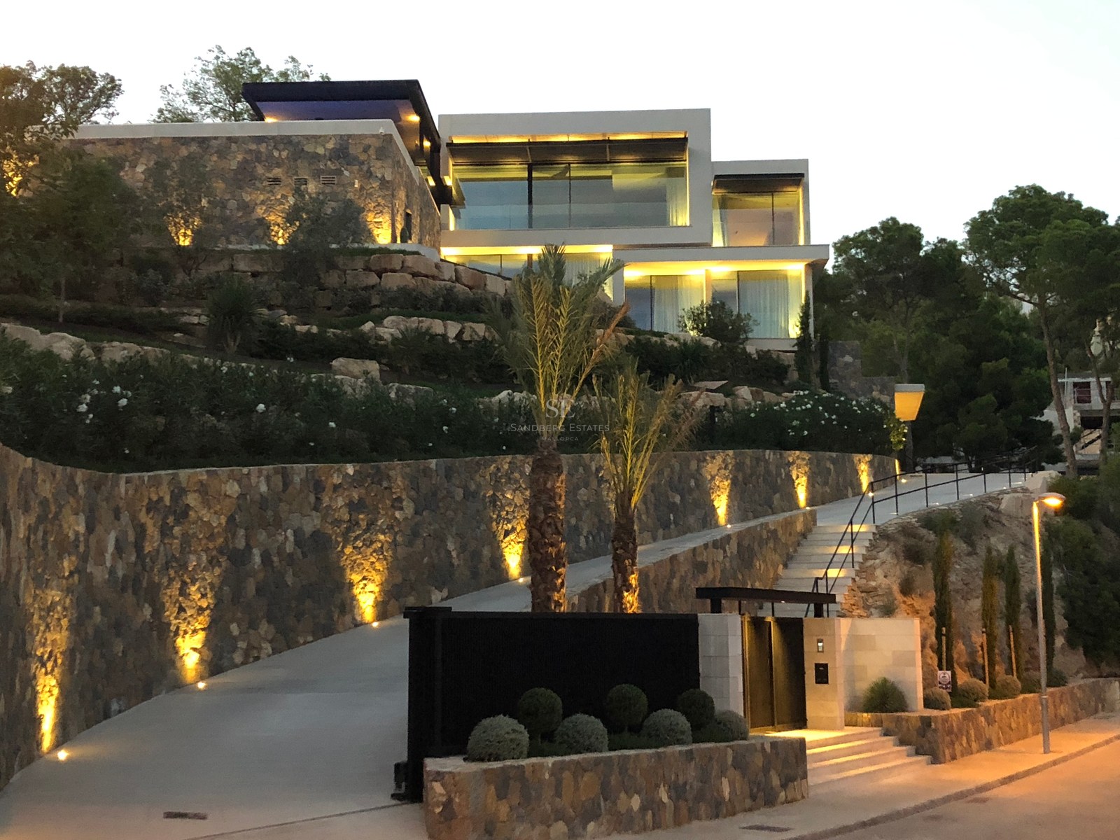 Architektonische Abendaufnahme einer terrassierten Luxusvilla mit Steinmauern und beleuchteter Einfahrt.