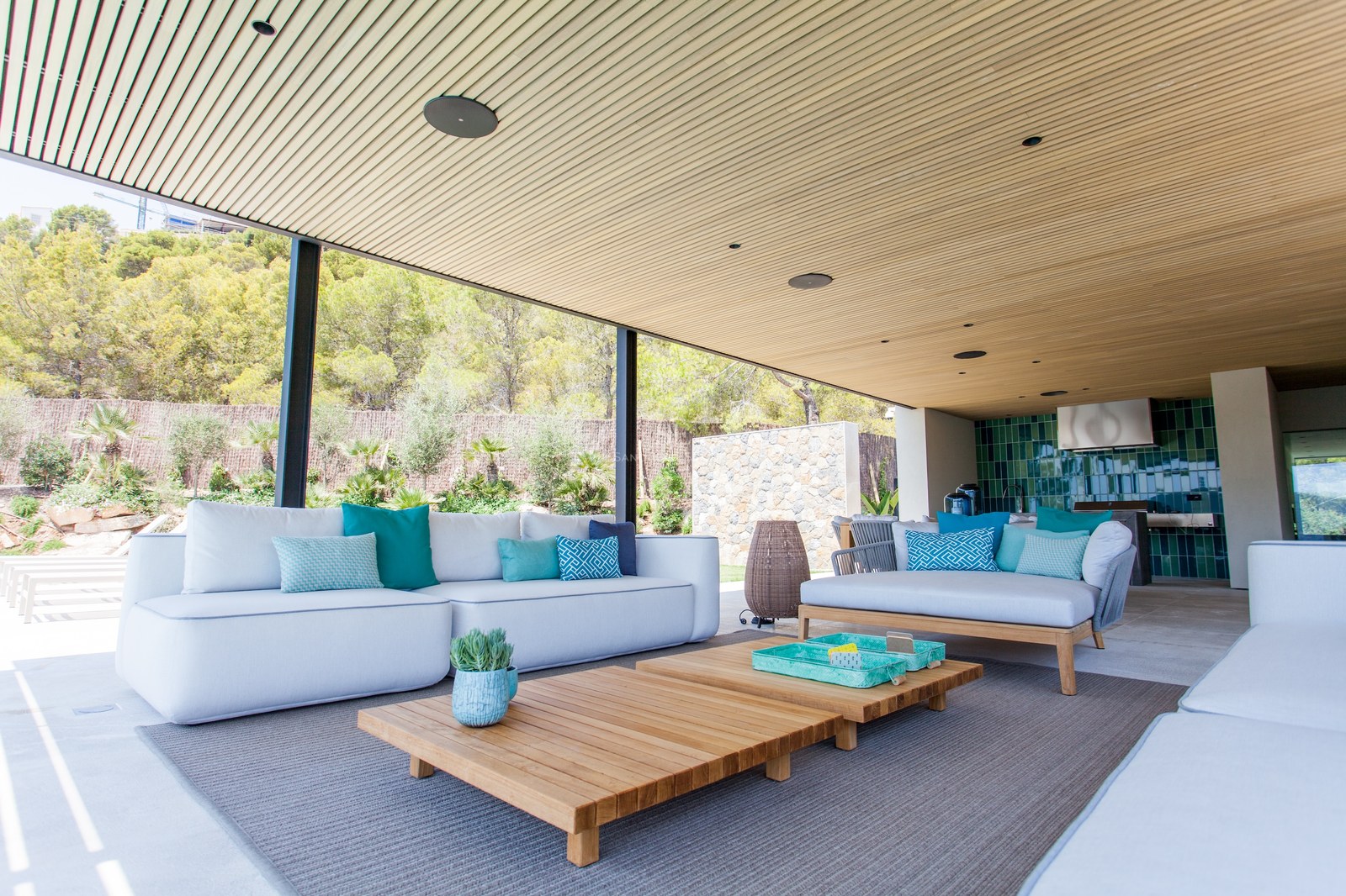 Moderne überdachte Terrasse mit weißen Sofas, türkisfarbenen Kissen, Holztischen und einer Sommerküche.