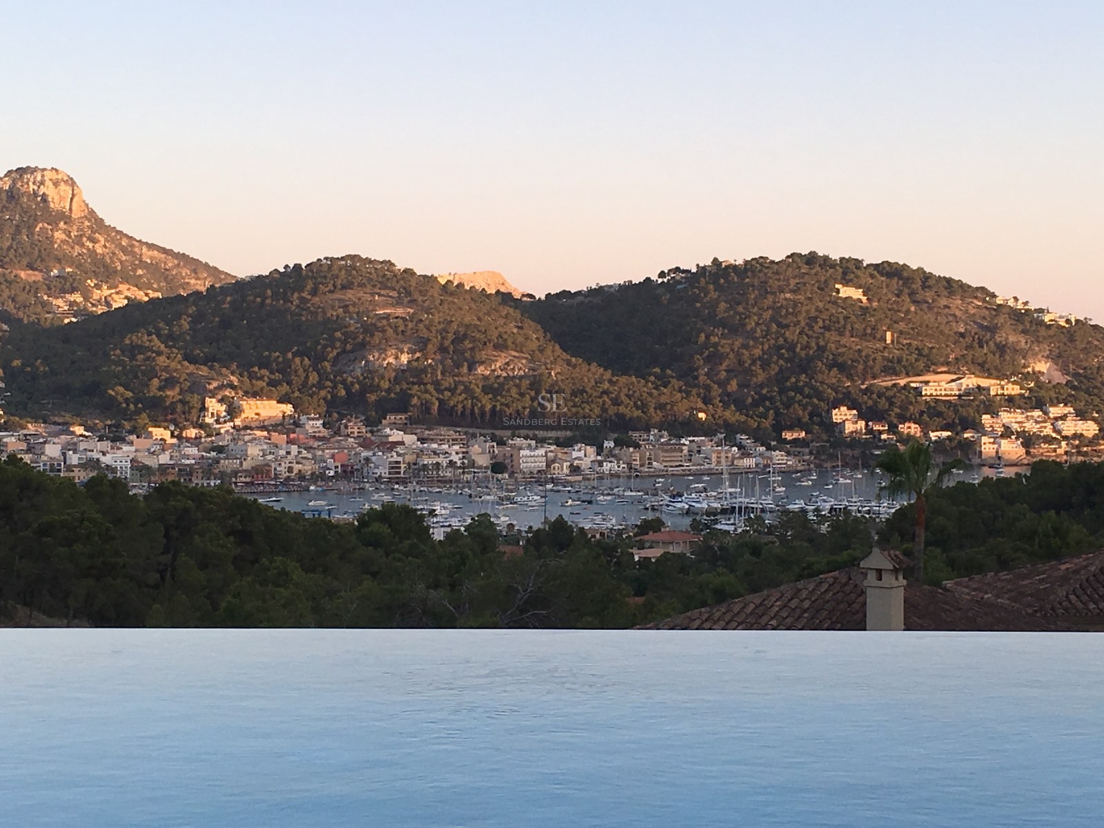 Rand eines Infinity-Pools mit Blick auf einen mediterranen Hafen mit Booten und grünen Bergen bei Sonnenuntergang.