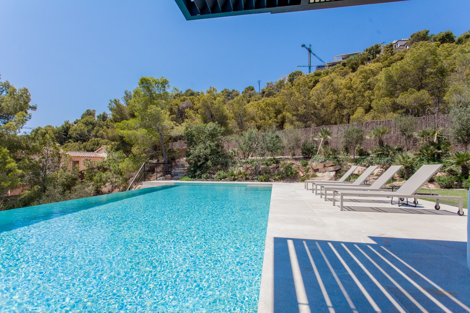 Großer Infinity-Pool mit türkisblauem Wasser und Steinterrasse mit grauen Liegen vor einem bewaldeten Hügel.