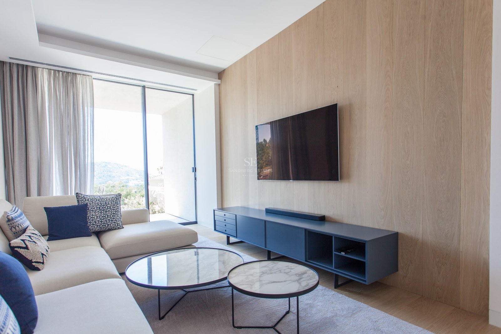 Modernes Wohnzimmer mit eichengetäfelter Wand, Wand-TV, Marmortischen und großen Fenstern mit Bergblick.