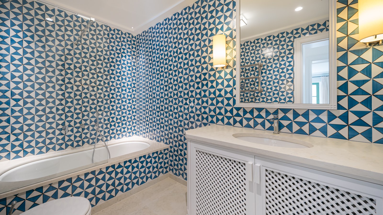 Baño con azulejos geométricos en azul y blanco, lavabo blanco con puertas de celosía y bañera integrada.