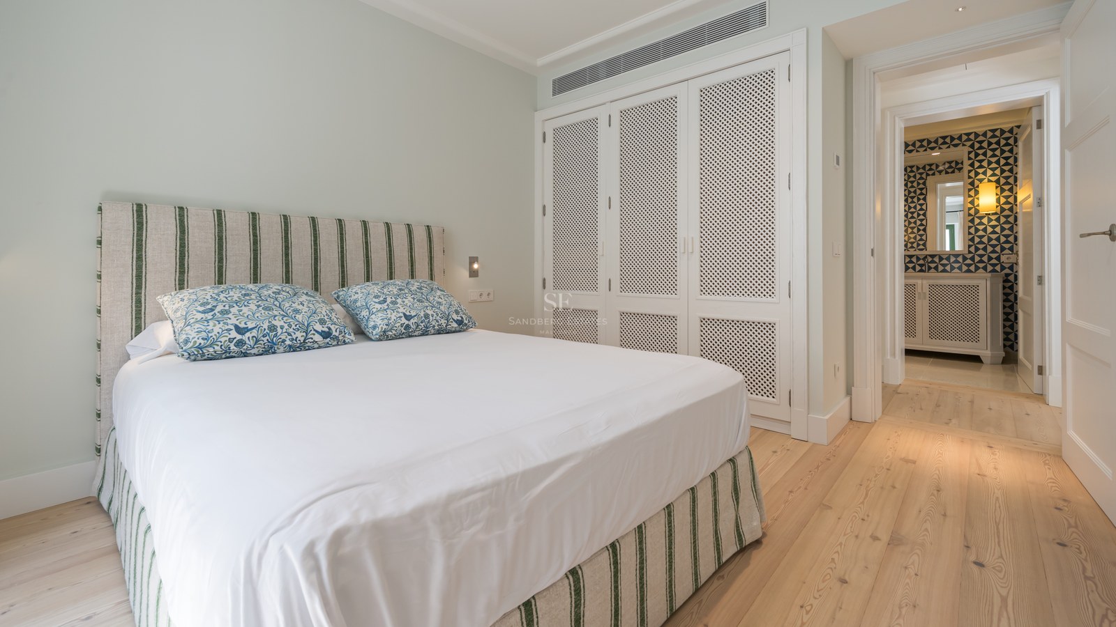 Dormitorio luminoso con cama blanca, cabecero de rayas verdes, suelos de madera clara y armarios empotrados blancos.