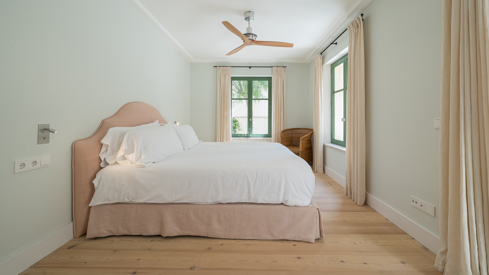 Dormitorio con cama blanca, cabecero rosa suave, ventilador de madera y ventanas de marco verde sobre suelo de roble.