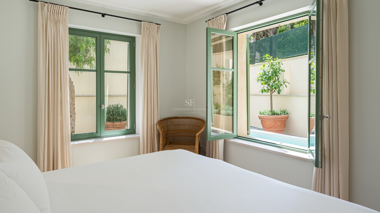 Dormitorio con cama blanca, ventanas de marco verde, sillón de mimbre y vistas a un patio con plantas.
