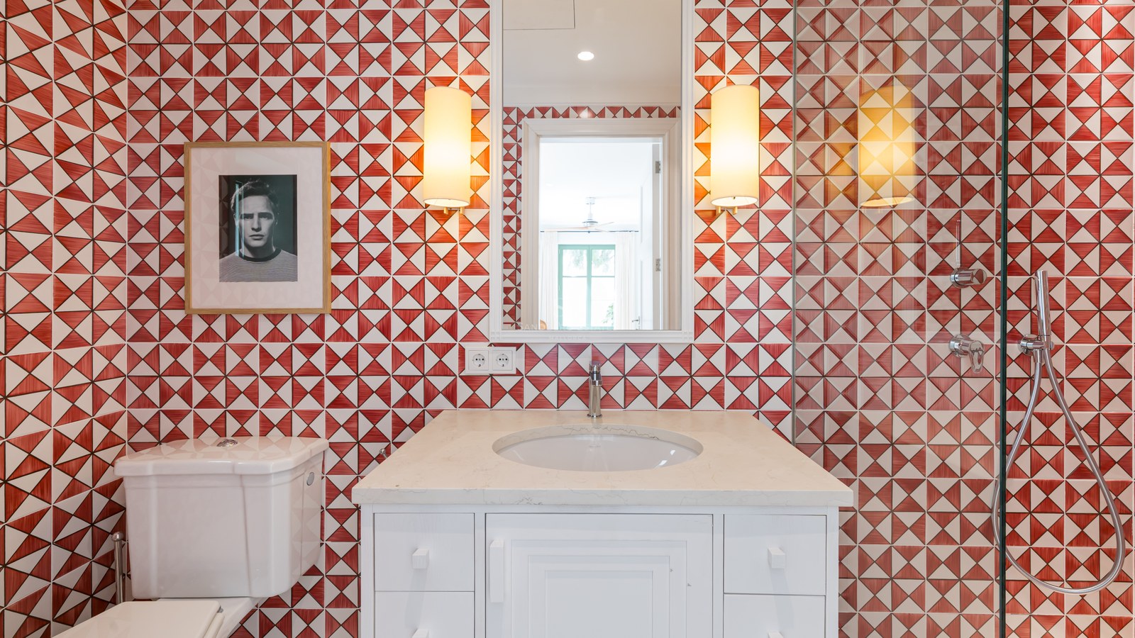Lavabo blanco con pared de azulejos geométricos rojos y blancos, espejo y apliques de pared.