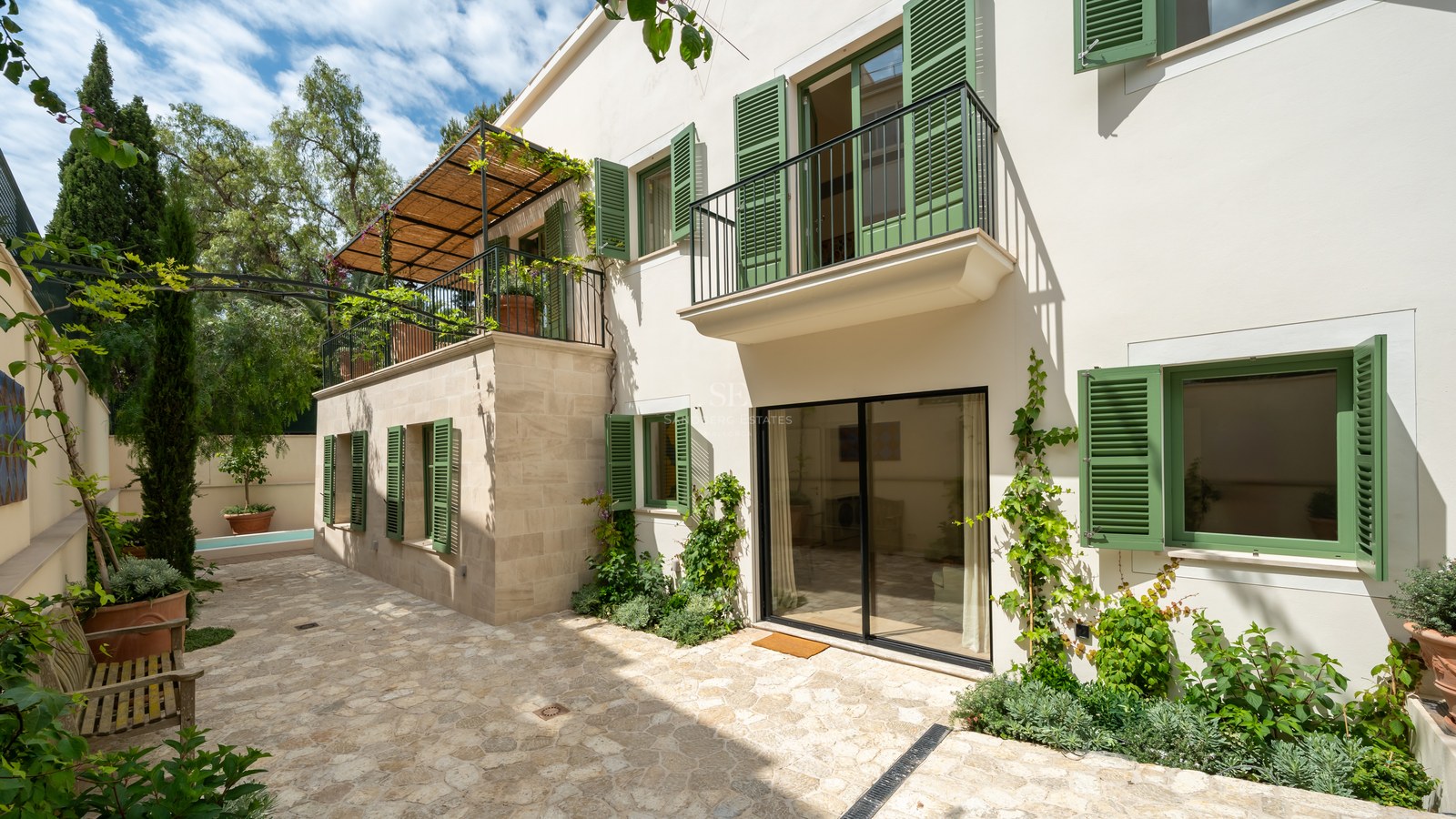 Fachada de villa blanca con persianas verdes, balcones de forja y patio de piedra natural bajo el sol.