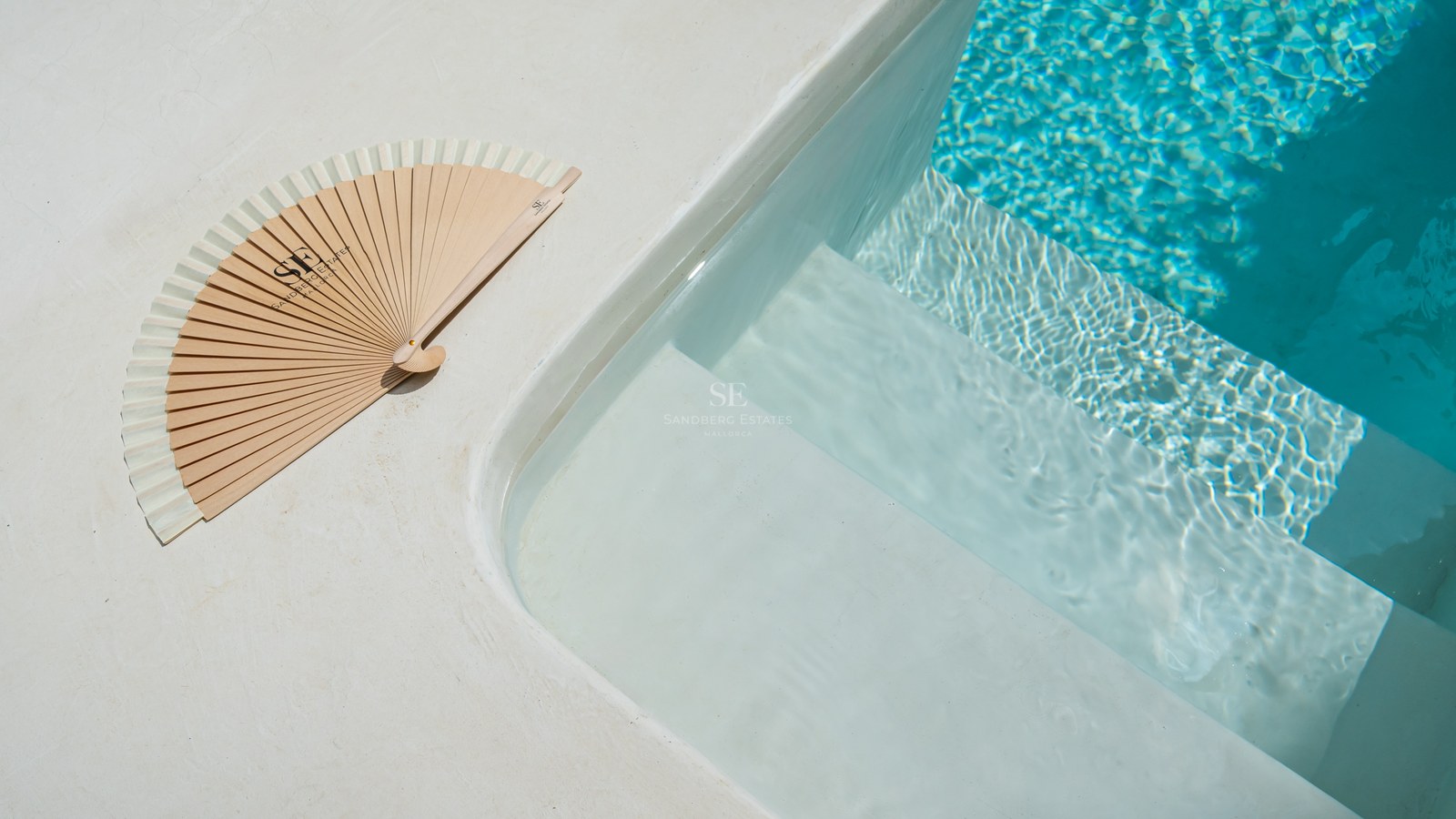 Primer plano de escalones blancos de piscina que entran en agua turquesa con un abanico de madera en el borde.
