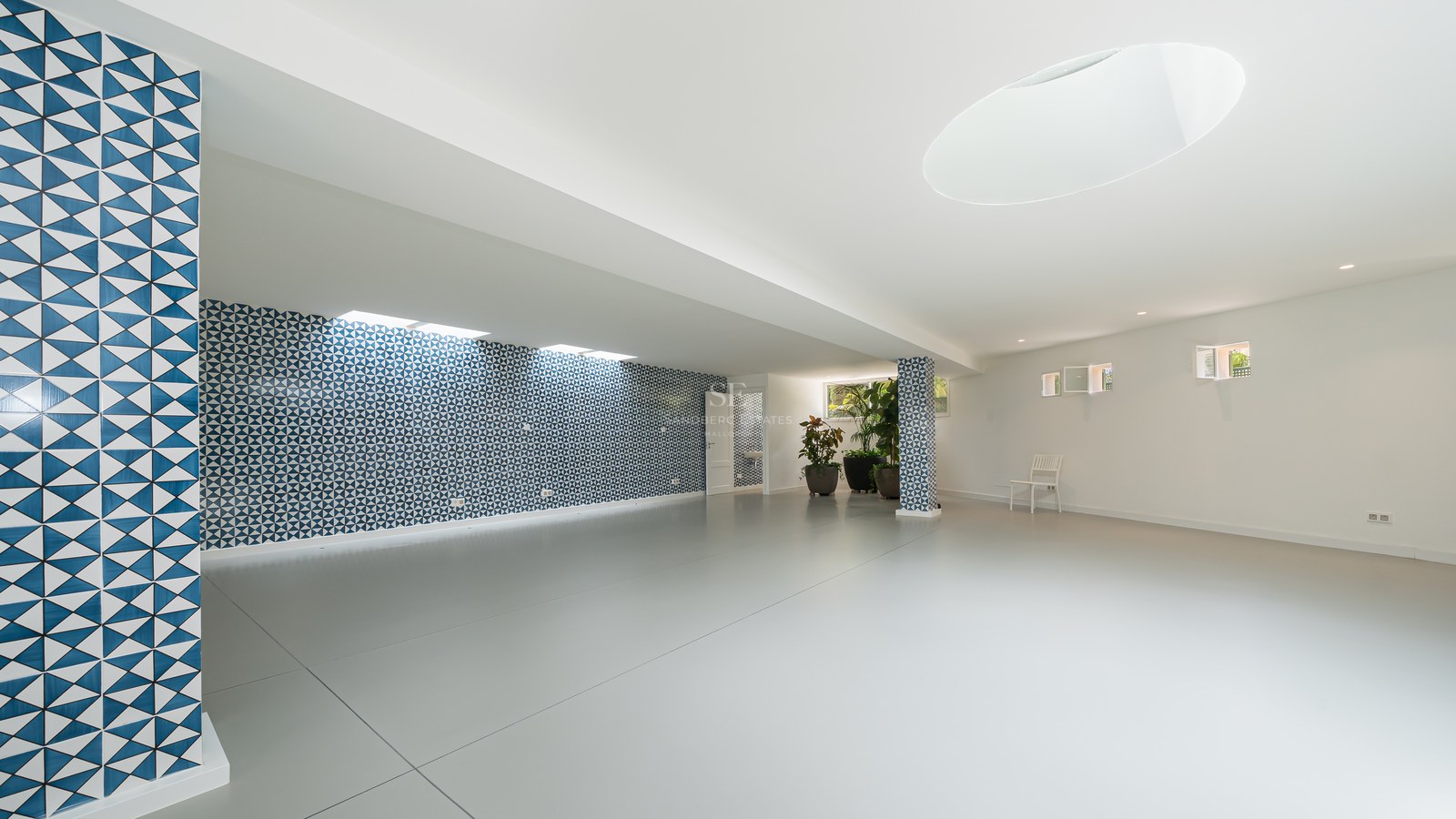 Amplia sala vacía con suelos de gres gris, pared de azulejos con motivos azules y plantas ornamentales.