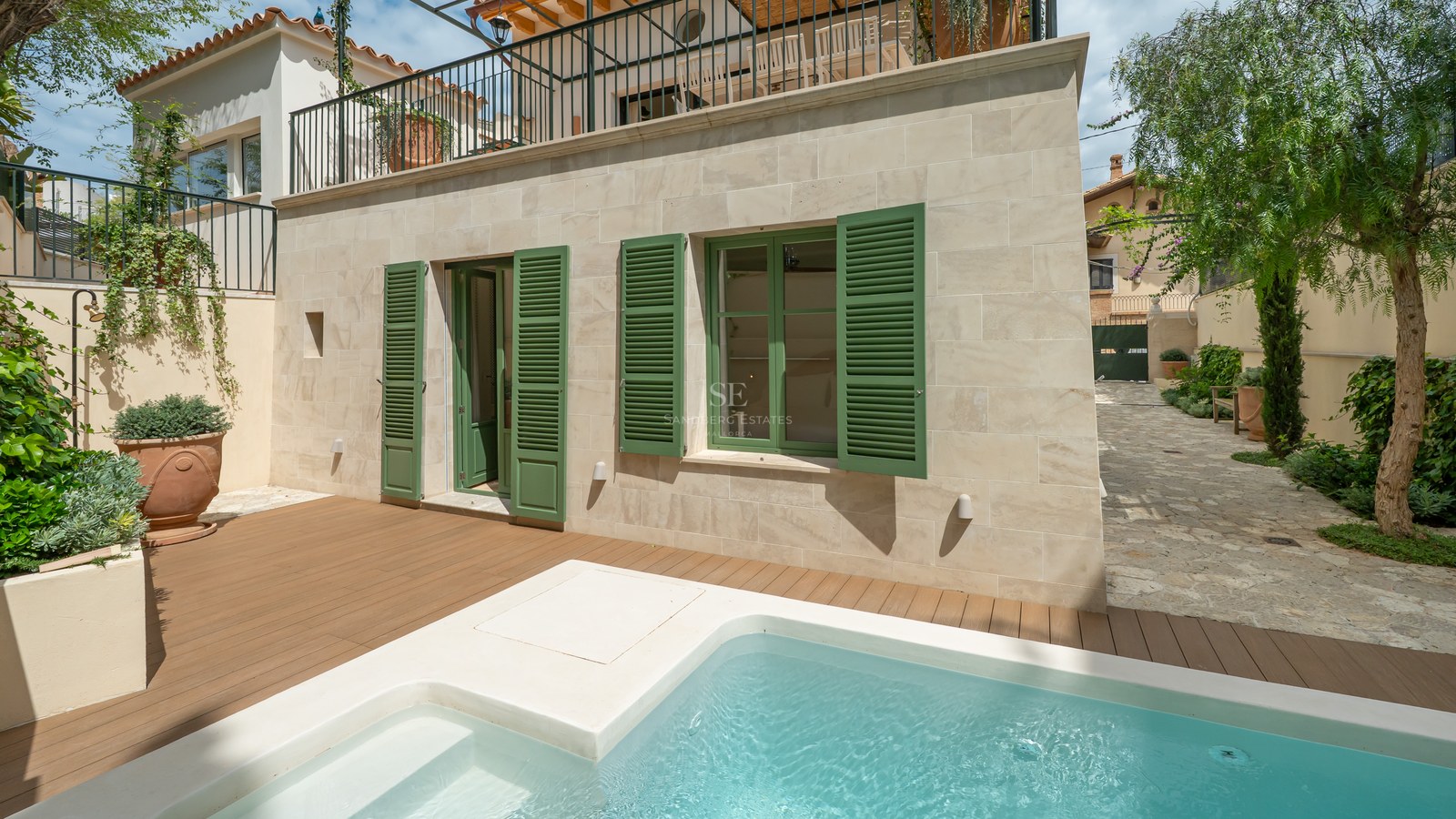 Piscina de inmersión moderna con tarima de madera junto a una fachada de piedra con contraventanas verdes.