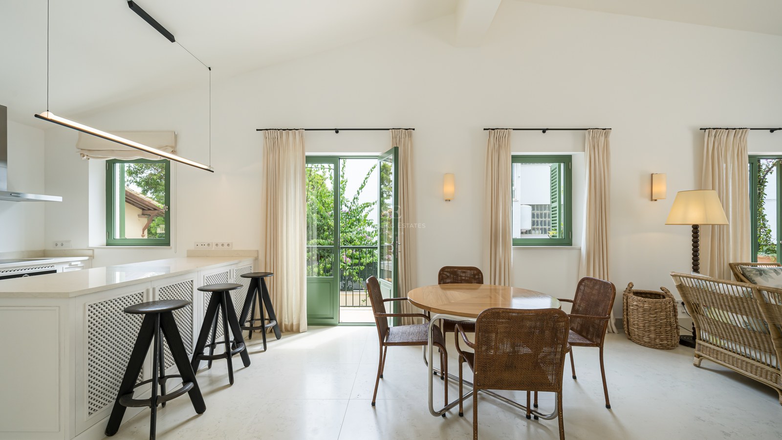 Comedor moderno con mesa redonda de madera, sillas de mimbre y puertas francesas verdes hacia un jardín frondoso.