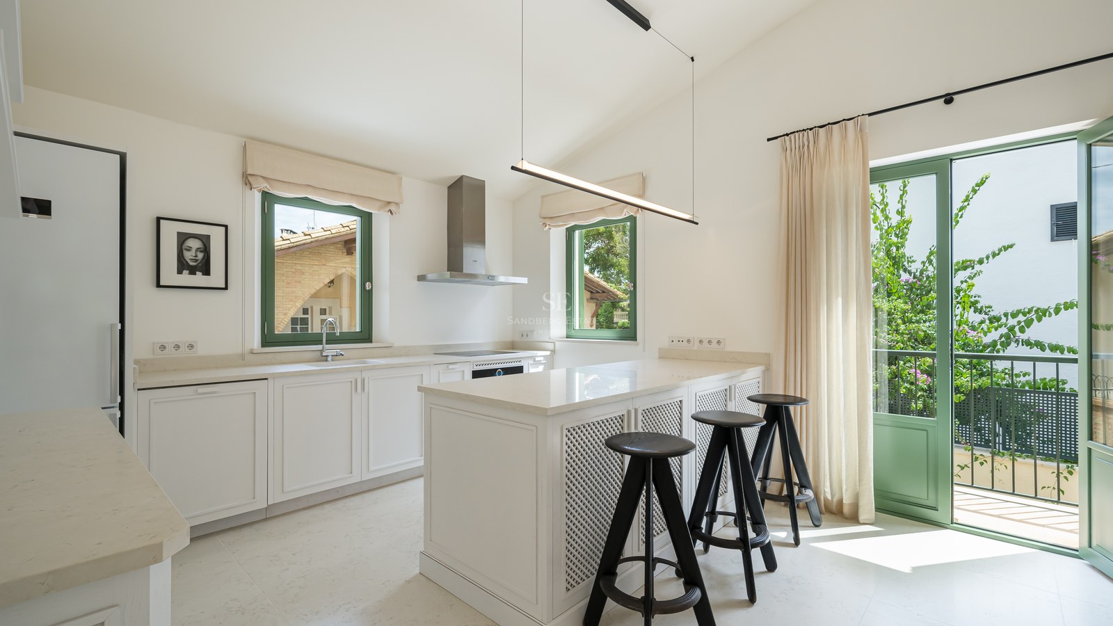 Cocina moderna blanca con isla central, taburetes negros y ventanas de marco verde inundadas de luz natural.