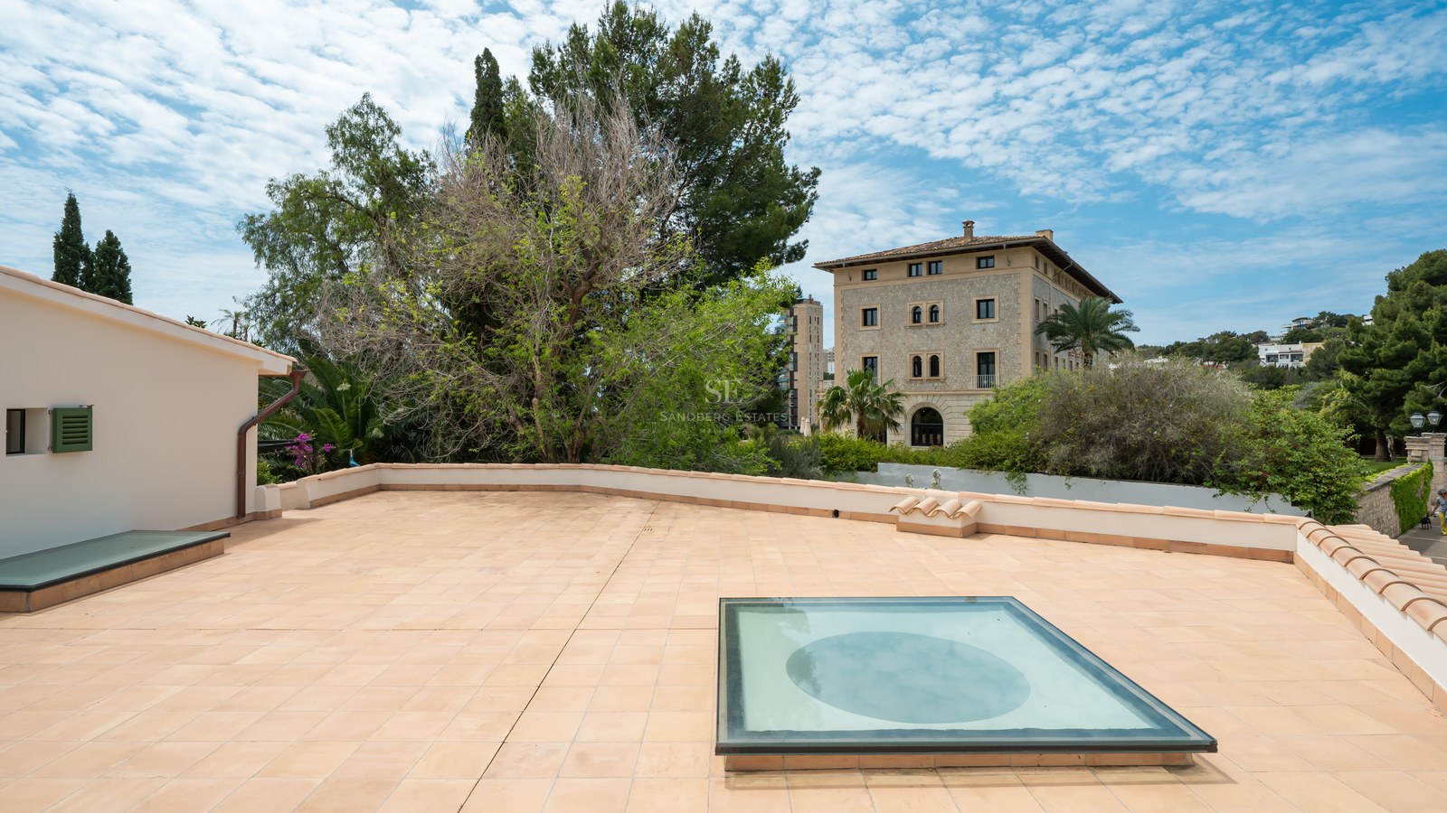 Gran terraza en la azotea con baldosas de terracota, claraboya de cristal y vistas a pinos y una mansión clásica.