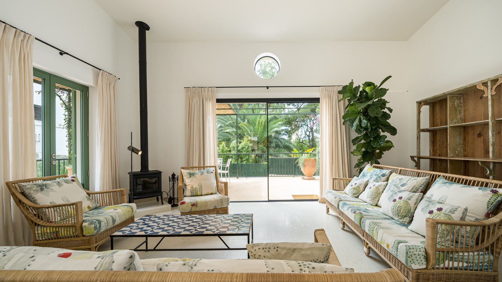 Salón soleado con muebles de bambú, estufa de leña negra y grandes puertas de cristal hacia un jardín verde.