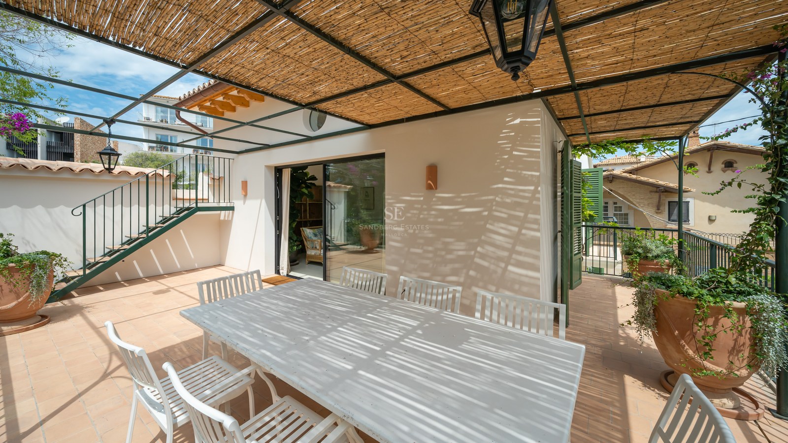 Mesa y sillas de madera blanca bajo una pérgola de cañizo con un farol tradicional en una terraza soleada.