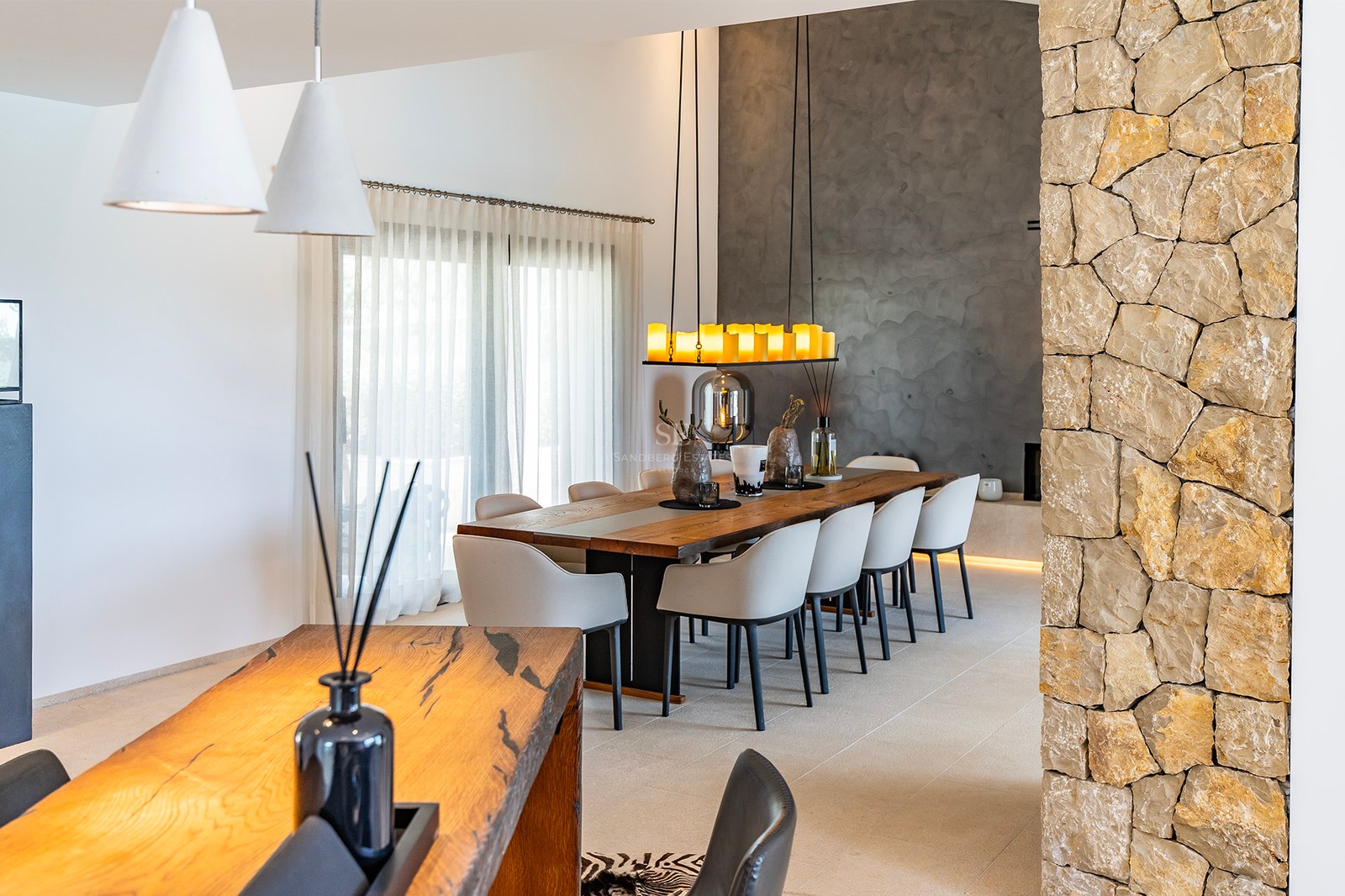 Modern matplats med långt massivt träbord, grå stolar, naturstensvägg och designad taklampa.
