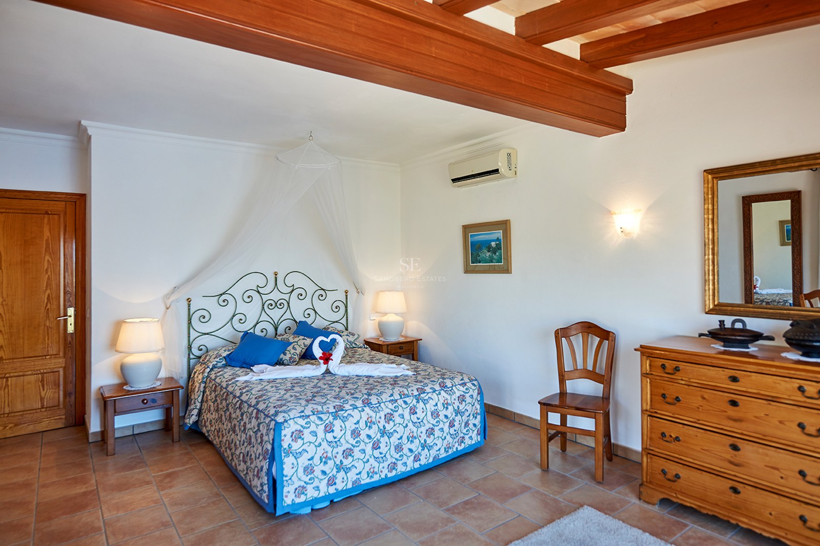 Schlafzimmer mit Terrakottaboden, sichtbaren Holzbalken, Schmiedeeisenbett und klassischen Holzmöbeln.