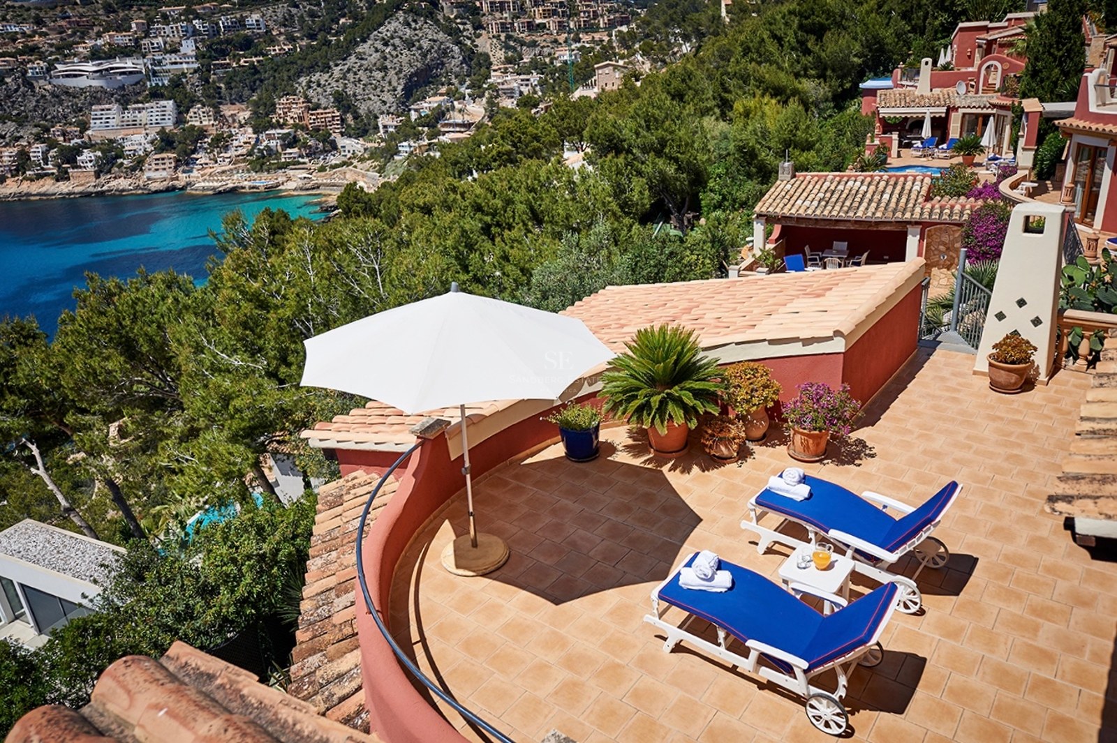 Sonnige Terrasse mit blauen Sonnenliegen, weißem Sonnenschirm und Panoramablick auf eine mediterrane Bucht.