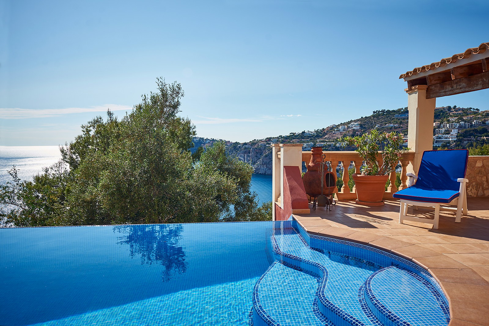 Infinity-Pool mit blauen Mosaikfliesen neben einer Terrakotta-Terrasse mit Blick auf das Mittelmeer und die Küste.