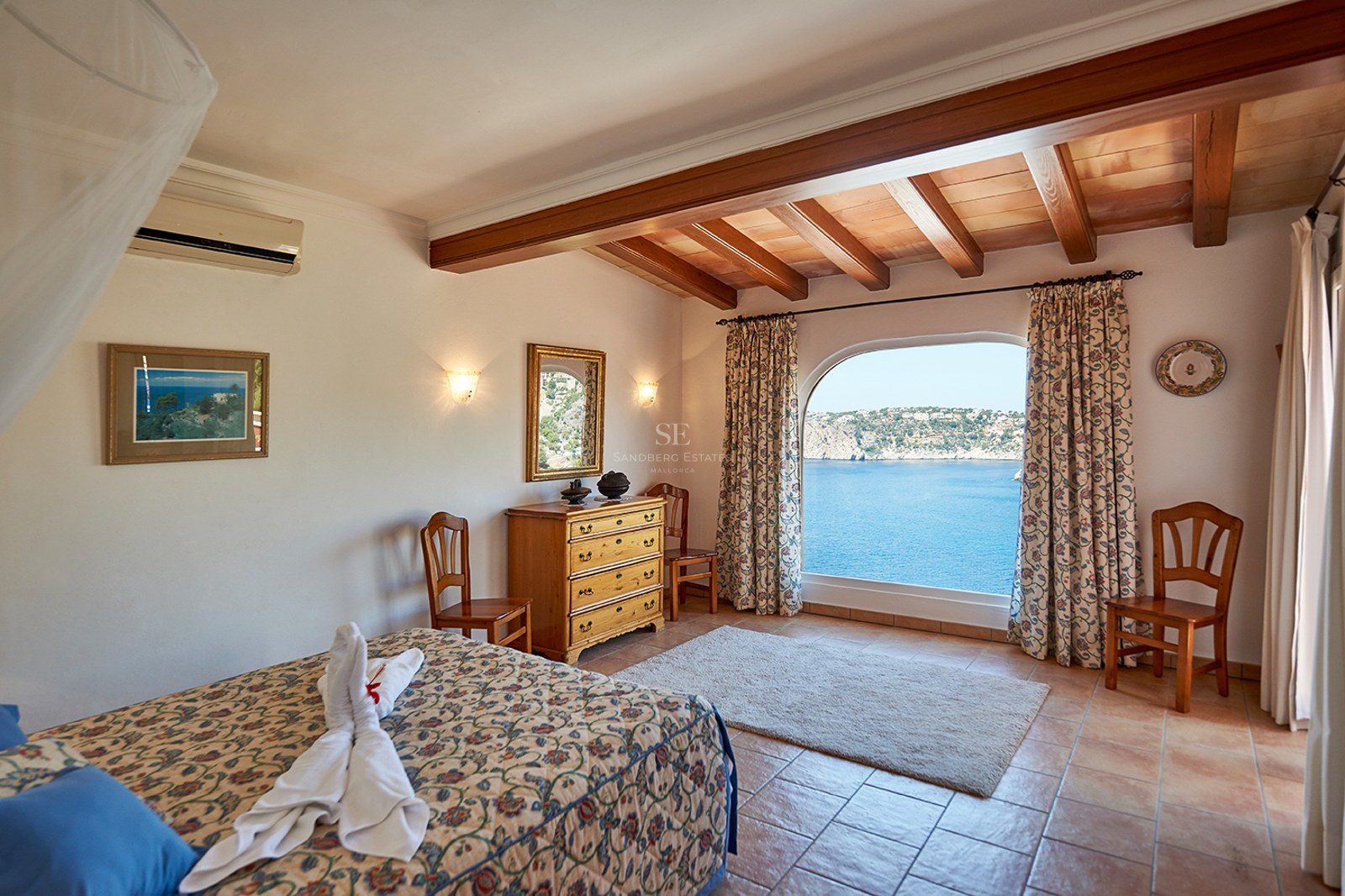 Schlafzimmer mit Terrakottaböden, Holzbalken und einem großen Bogenfenster mit Blick auf das blaue Meer.