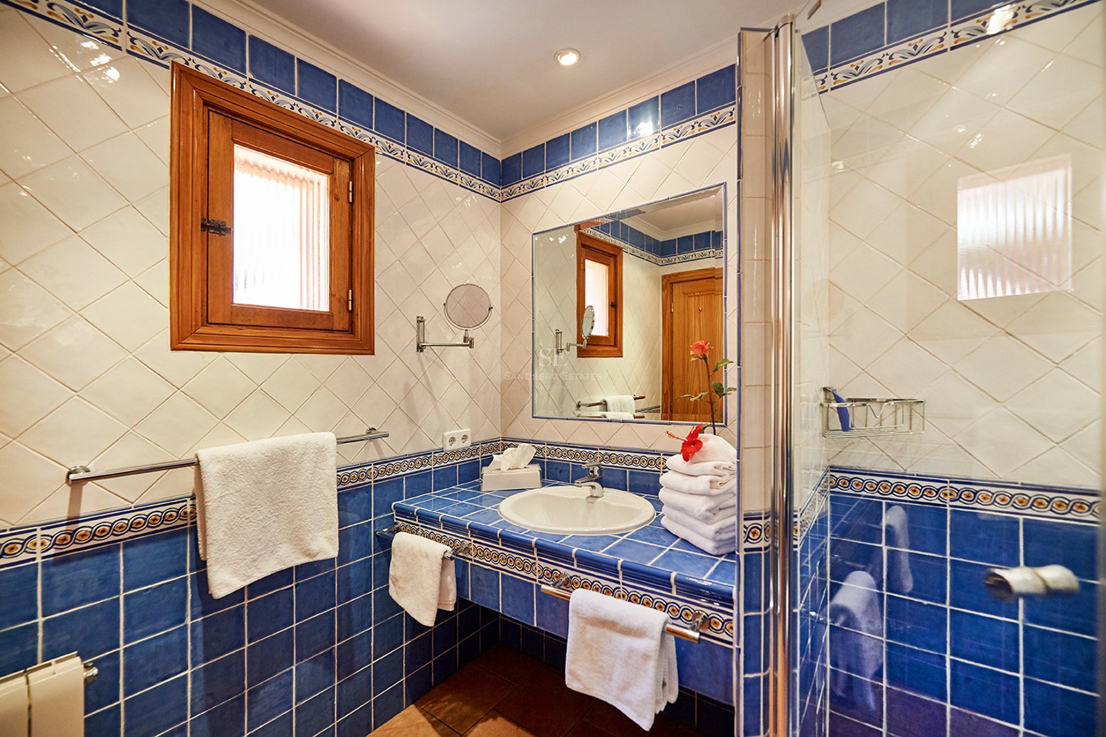 Badezimmer mit blau-weißen Fliesen, Waschbecken, Spiegel, Glasdusche und Holzfenster.