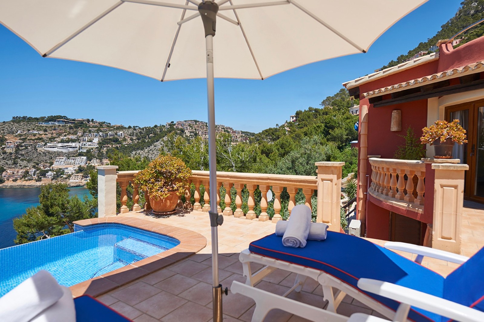 Sonnige Terrasse mit kleinem Pool, blauen Liegen und Sonnenschirm mit Blick auf eine Bucht und Hügel.