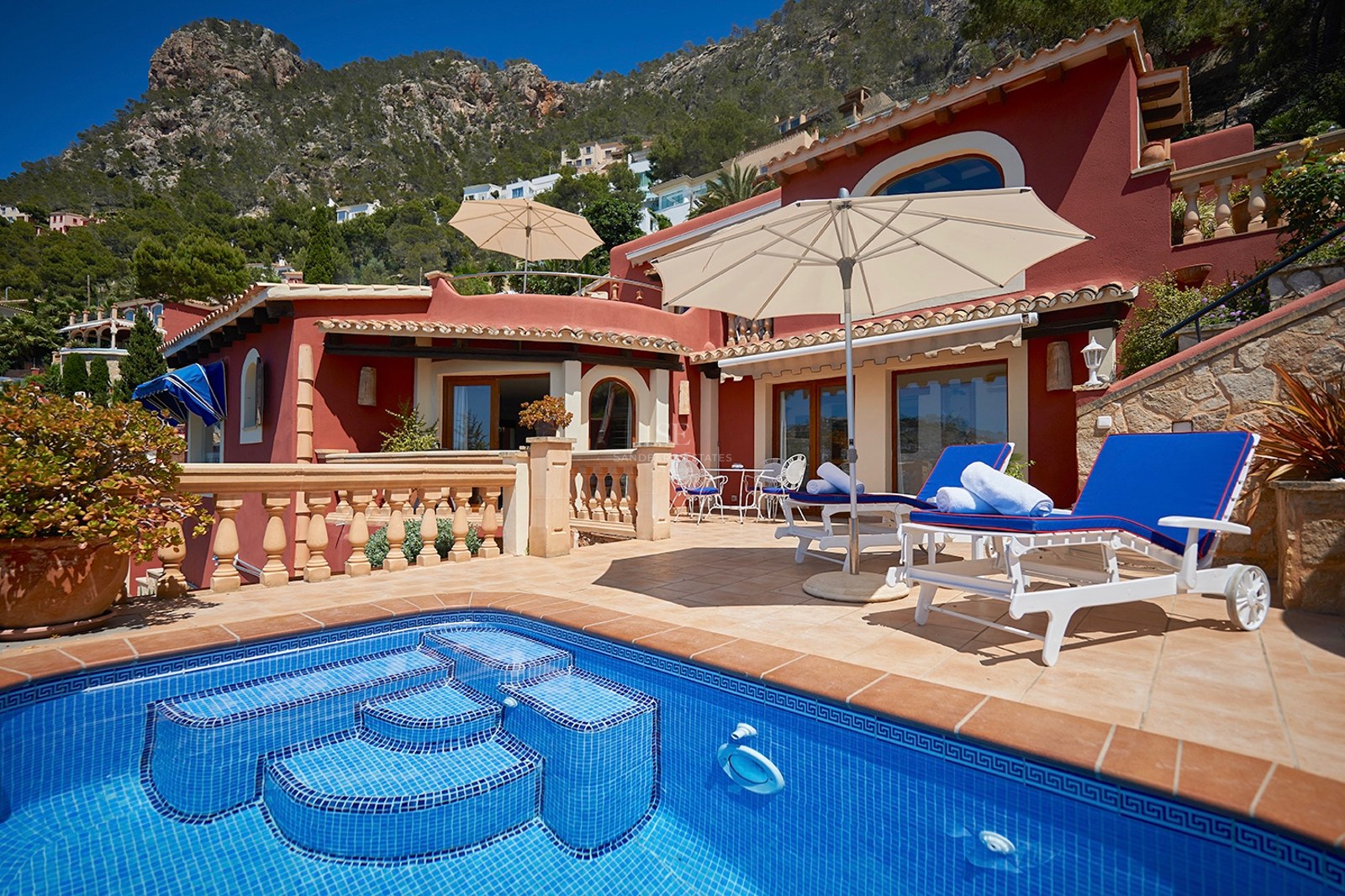 Blauer Pool und Sonnendeck mit Liegen vor einer terrakottafarbenen Villa mit Bergen im Hintergrund.