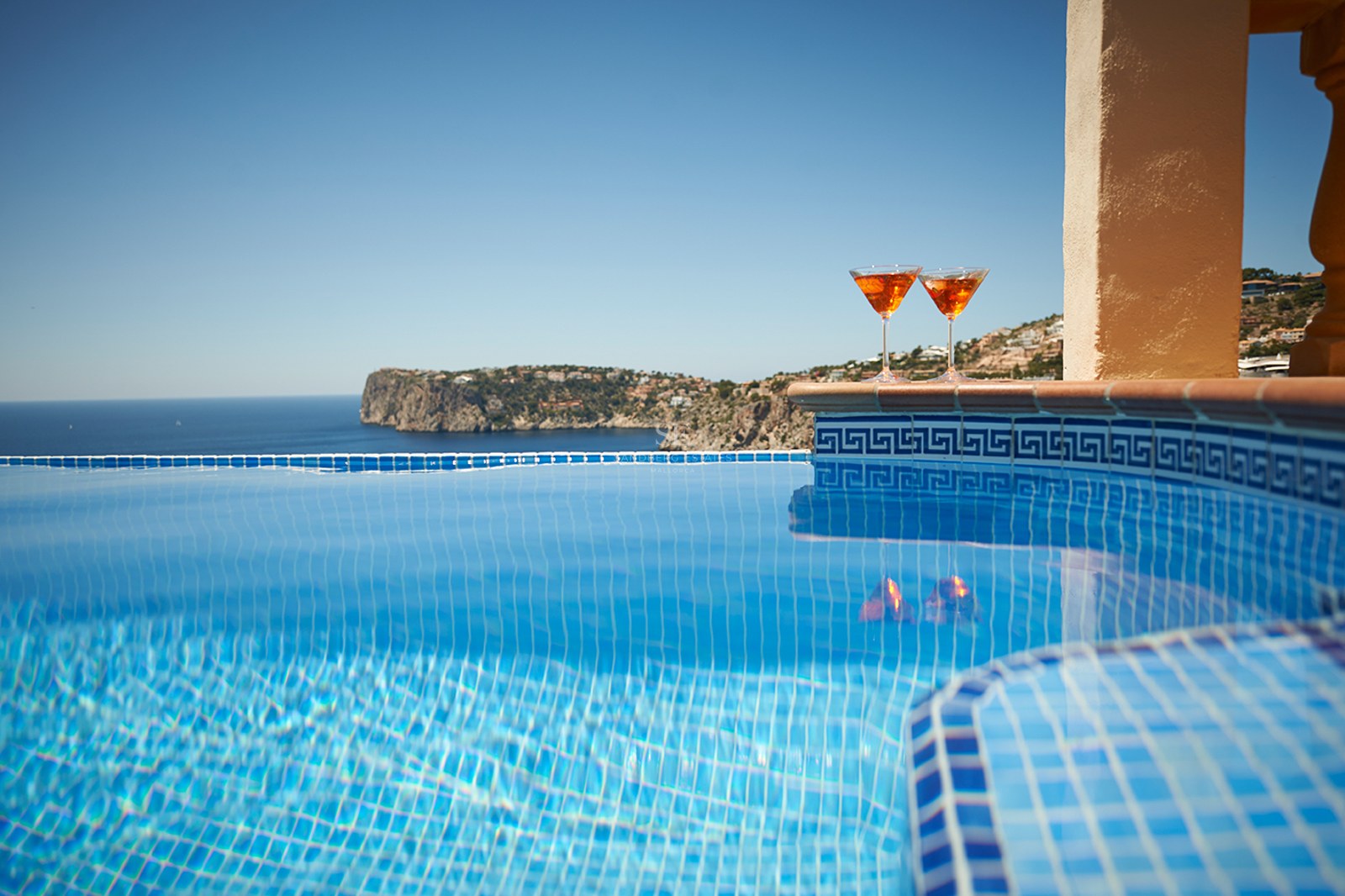 Nahaufnahme eines blau gefliesten Infinity-Pools mit zwei Cocktails am Rand und Blick auf das Meer.