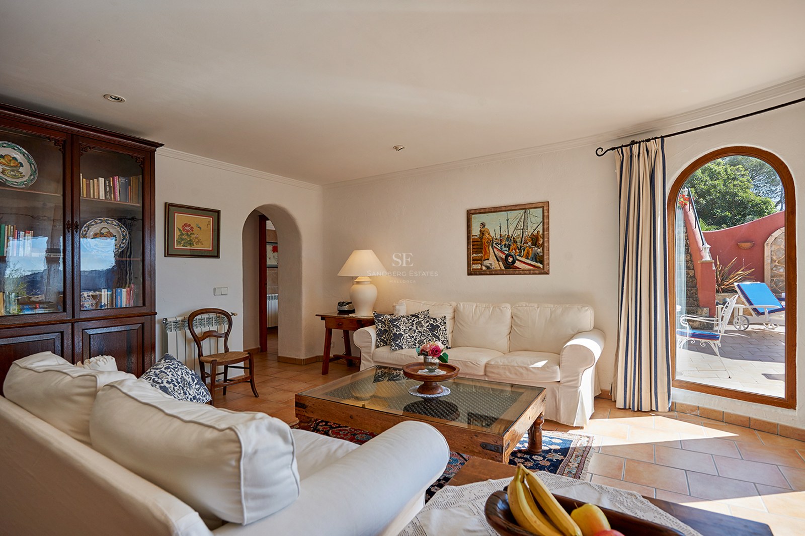 Helles Wohnzimmer mit weißen Sofas, dunklen Holzmöbeln, Terrakottafliesen und einer Bogentür zur Terrasse.