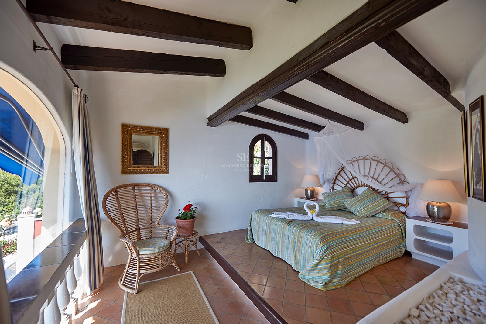 Helles Schlafzimmer mit dunklen Holzbalken, Weidenkopfteil, Terrakottaboden und Rundbogenfenster.