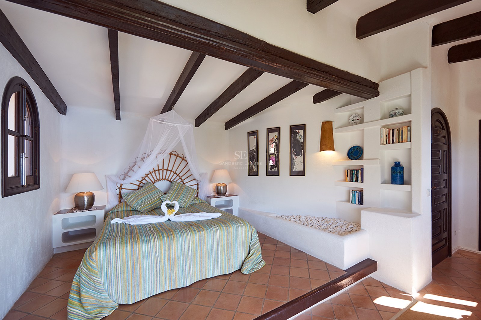 Schlafzimmer mit Korbgeflecht-Bett, dunklen Holzbalken, Terrakottafliesen und weißen Wänden mit eingebauten Regalen.
