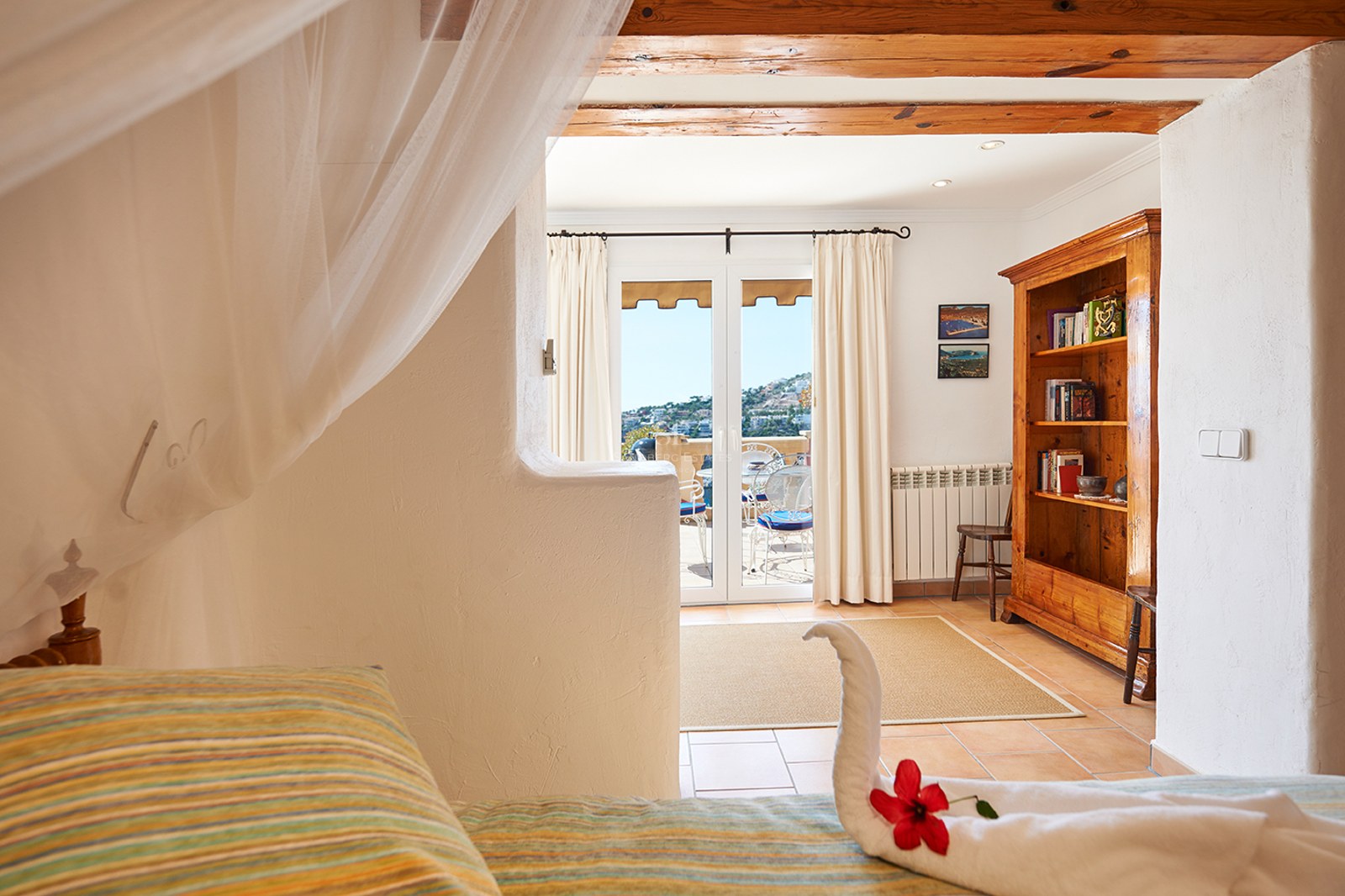 Rustikales Schlafzimmer mit Holzbalken, Himmelbett mit Handtuch-Schwan und Türen zu einer sonnigen Terrasse.