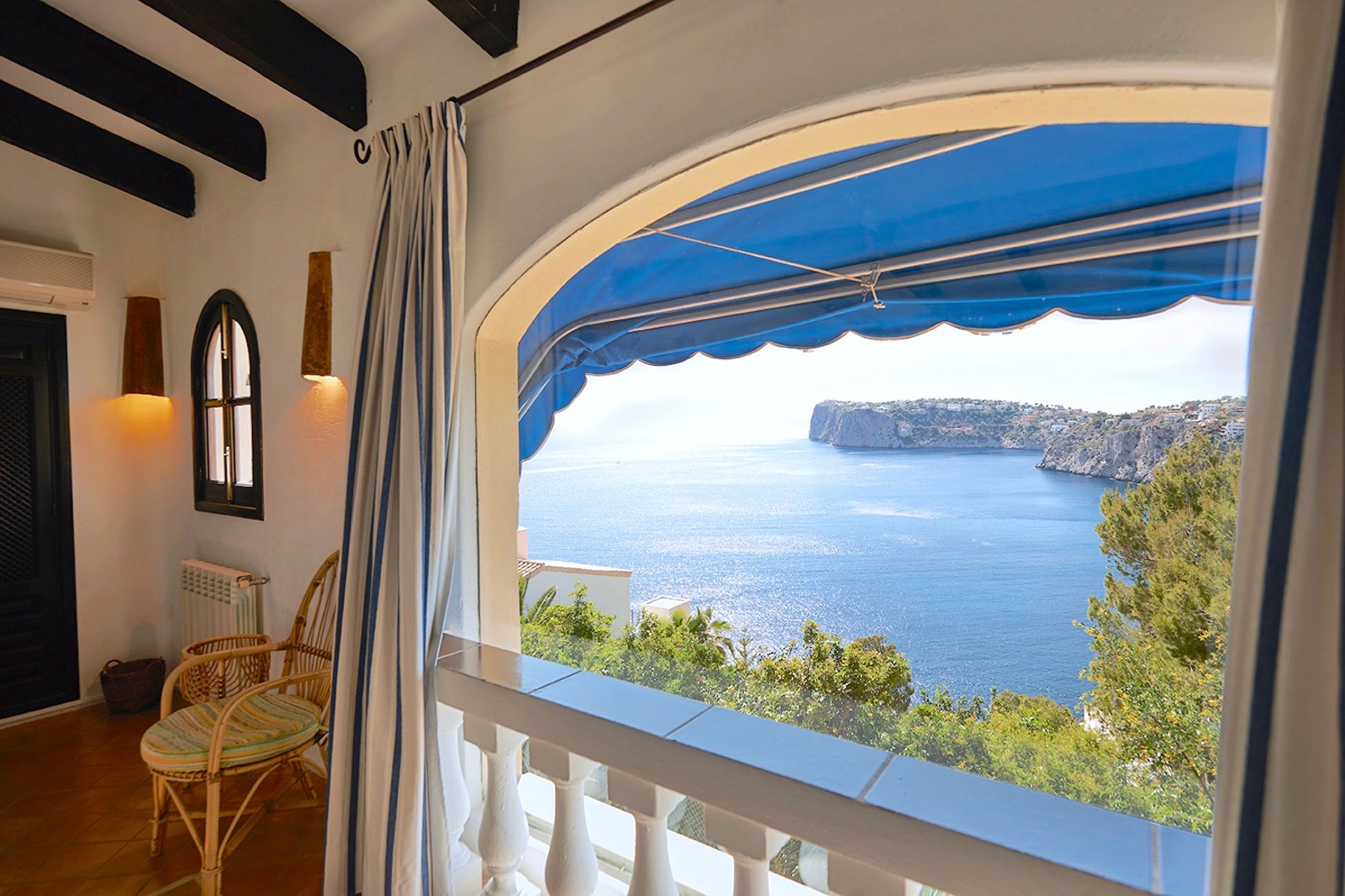 Überdachte Terrasse mit Korbsessel und blauer Markise, mit Blick auf das blaue Meer und Klippen.