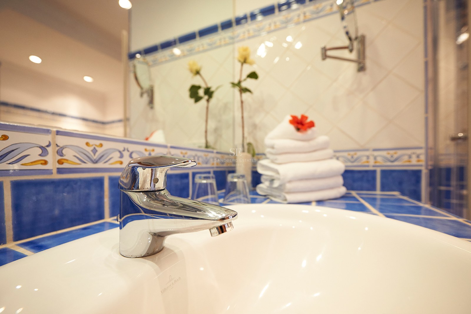 Nahaufnahme eines weißen Porzellanwaschbeckens und verchromten Wasserhahns mit blau gemusterten mediterranen Fliesen.