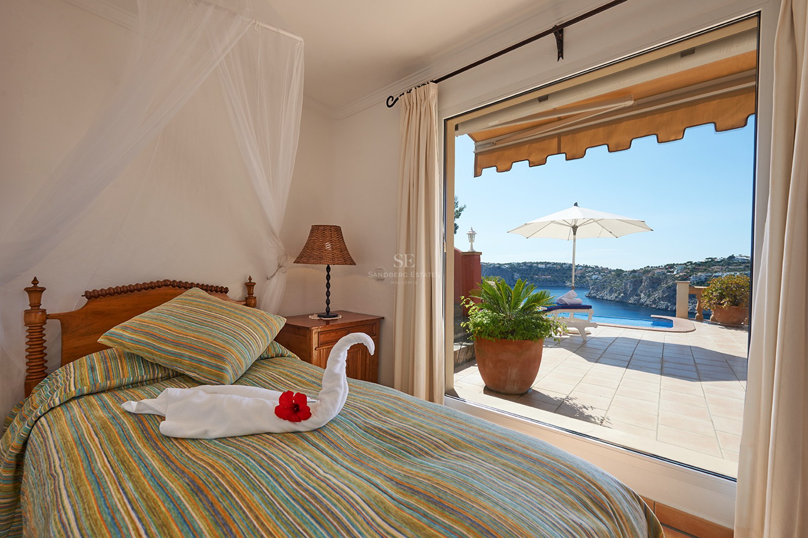 Schlafzimmer mit gestreiftem Bett und Handtuchschwan mit Blick auf eine Terrasse mit Pool und blauem Meer.