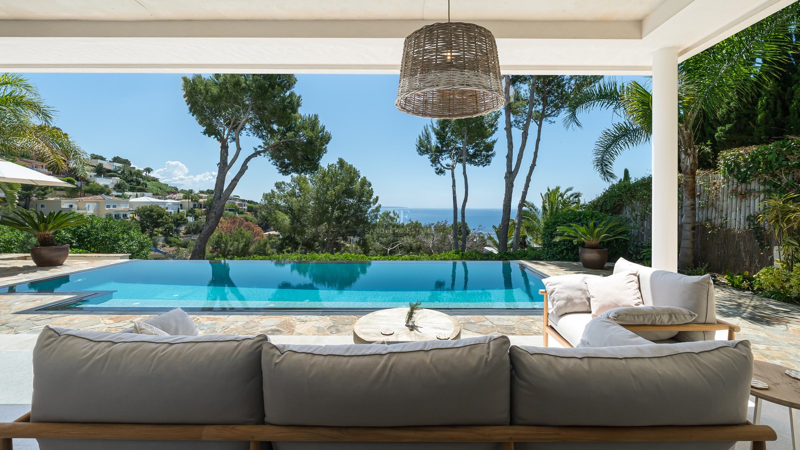 Infinitypool och loungegrupp under en skuggad terrass med utsikt över havet och lummiga tallar.