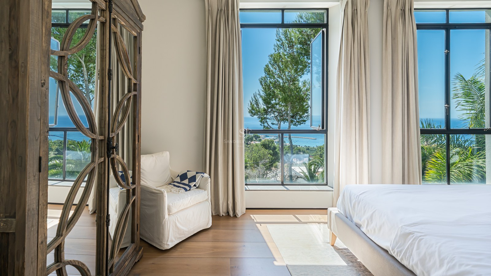 Ljust master bedroom med stora fönster mot havet, en vit fåtölj och en utsmyckad garderob i trä.