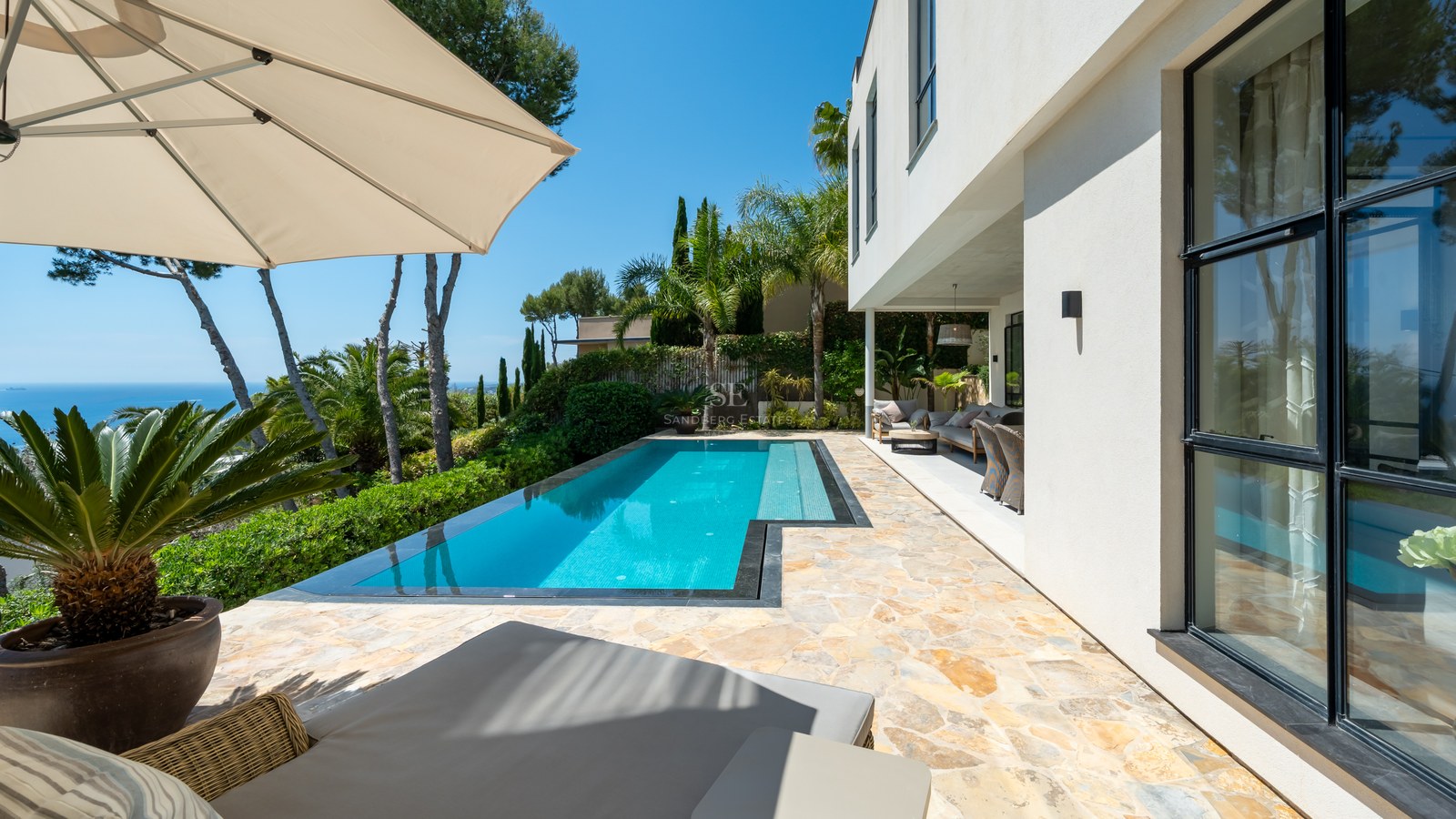En turkos pool på en stenterrass bredvid en vit modern villa med utsikt över havet.