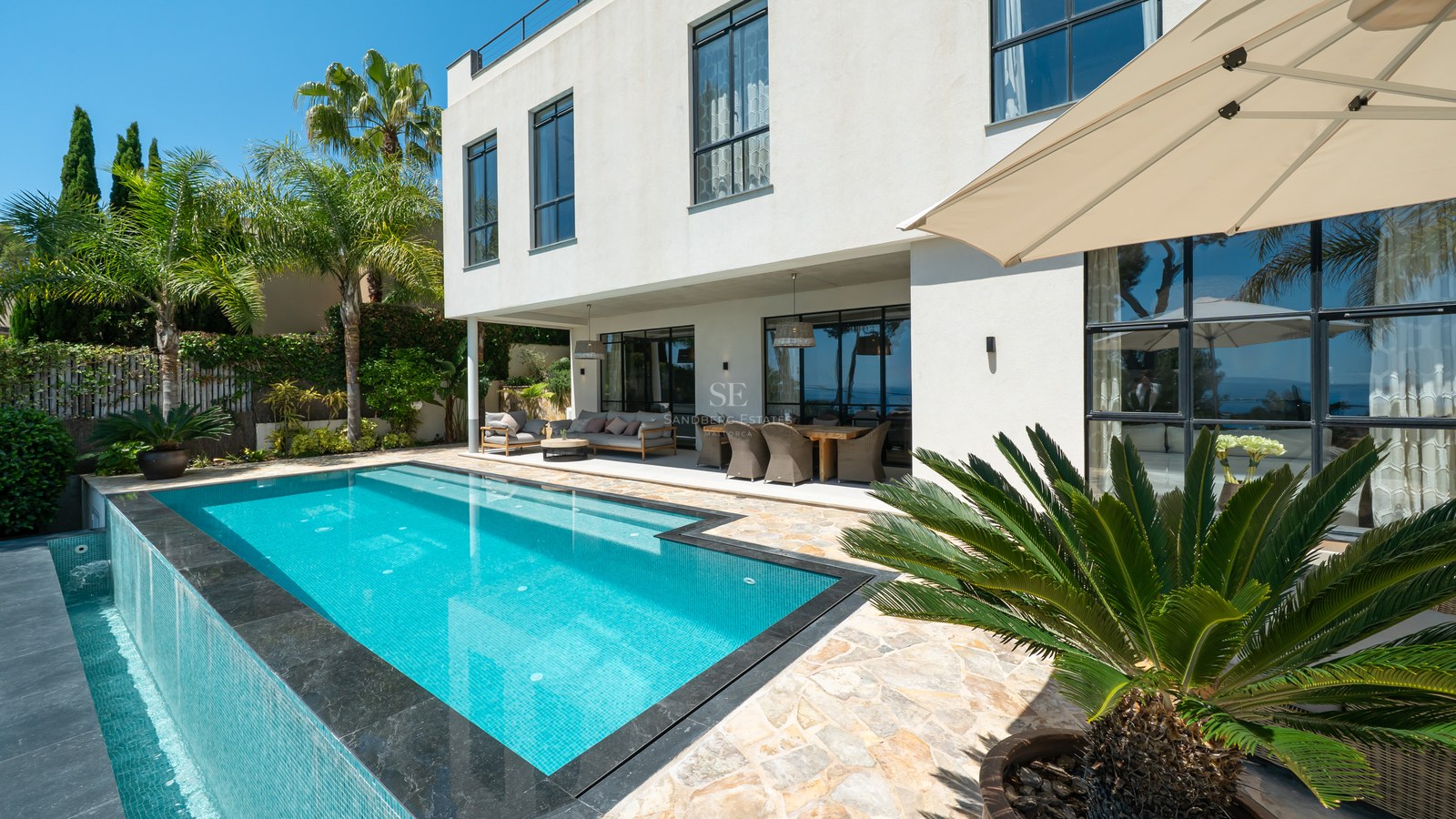 Ljus utomhusscen med turkos pool, täckt terrass och modern vit villa omgiven av palmer.