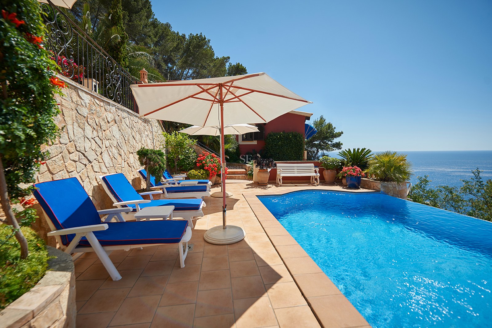 Solig poolterrass med blå solstolar och vita parasoller med utsikt över det glittrande Medelhavet.