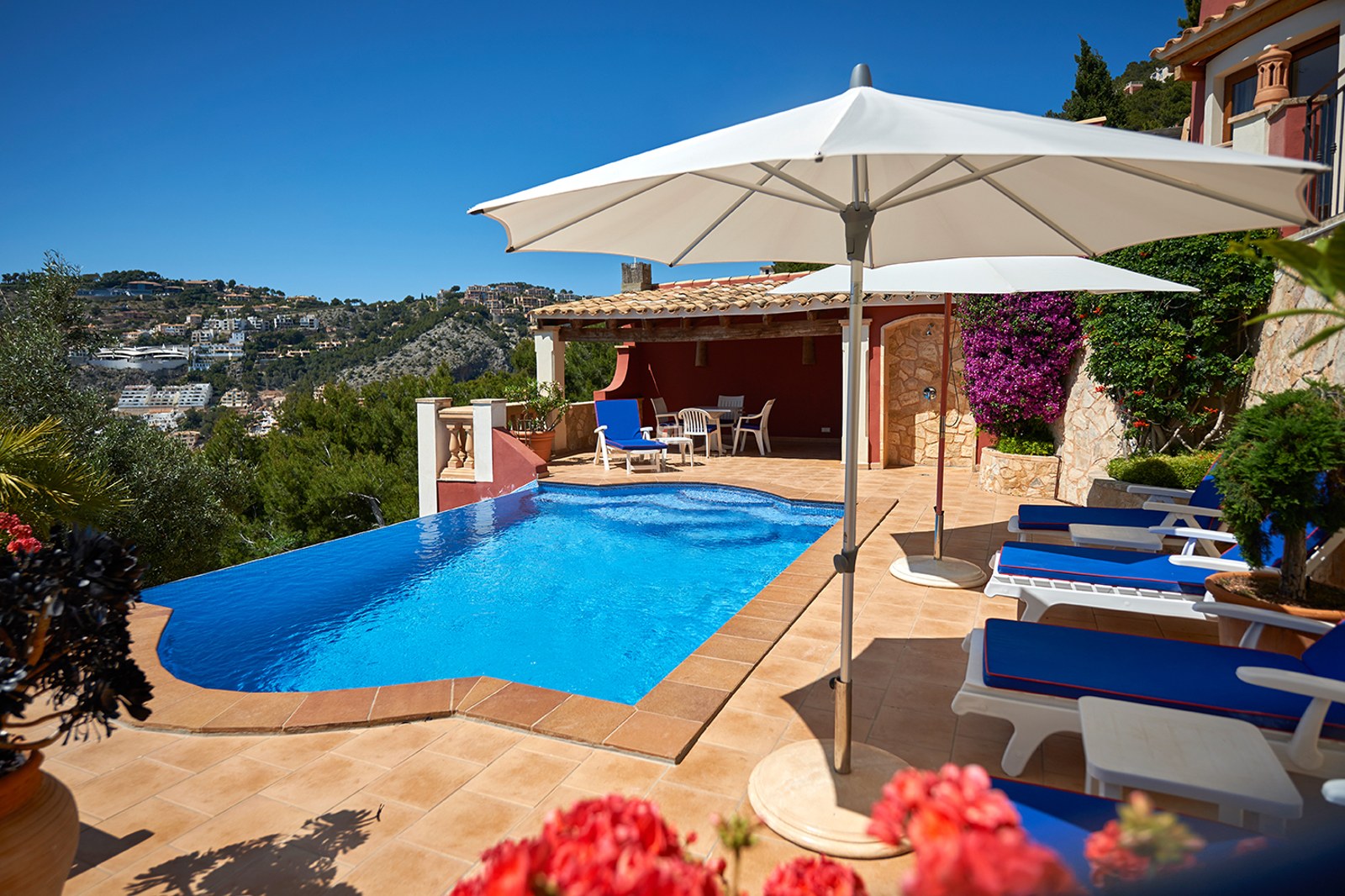 En klarblå pool på en terrass med terrakottaklinker, solstolar och parasoller med utsikt över kullar och havet.
